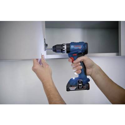 Une personne utilise une perceuse sans fil bleue pour installer ou resserrer une charnière sur une porte de placard blanche, illustrant une tâche de bricolage domestique.