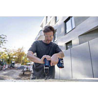 Ouvrier utilisant une perceuse électrique sur un chantier de construction, portant des lunettes de sécurité, avec un bâtiment et des arbres en arrière-plan.