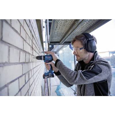 Une personne portant un équipement de protection utilise une perceuse sur un mur en brique, avec des échafaudages et des matériaux de construction en arrière-plan.