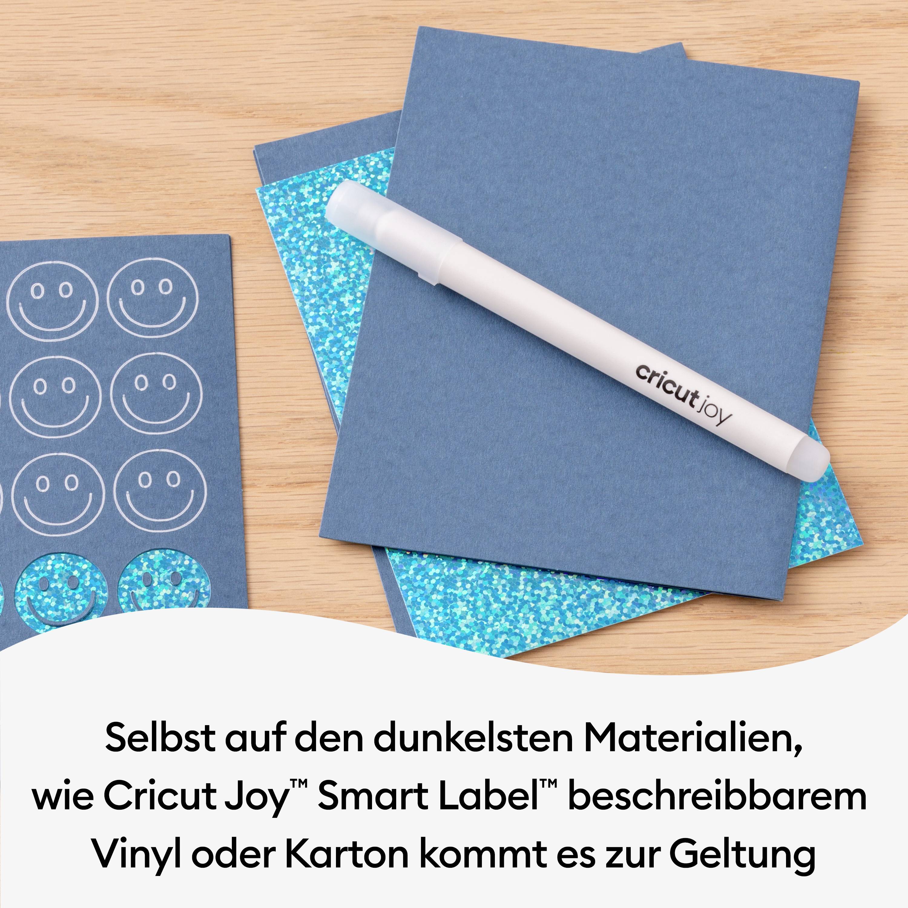 Un stylo Cricut Joy est posé sur des cartes bleues et scintillantes. À côté se trouvent des papiers avec des visages souriants. Le texte indique que le Cricut Joy fonctionne bien sur des matériaux sombres.