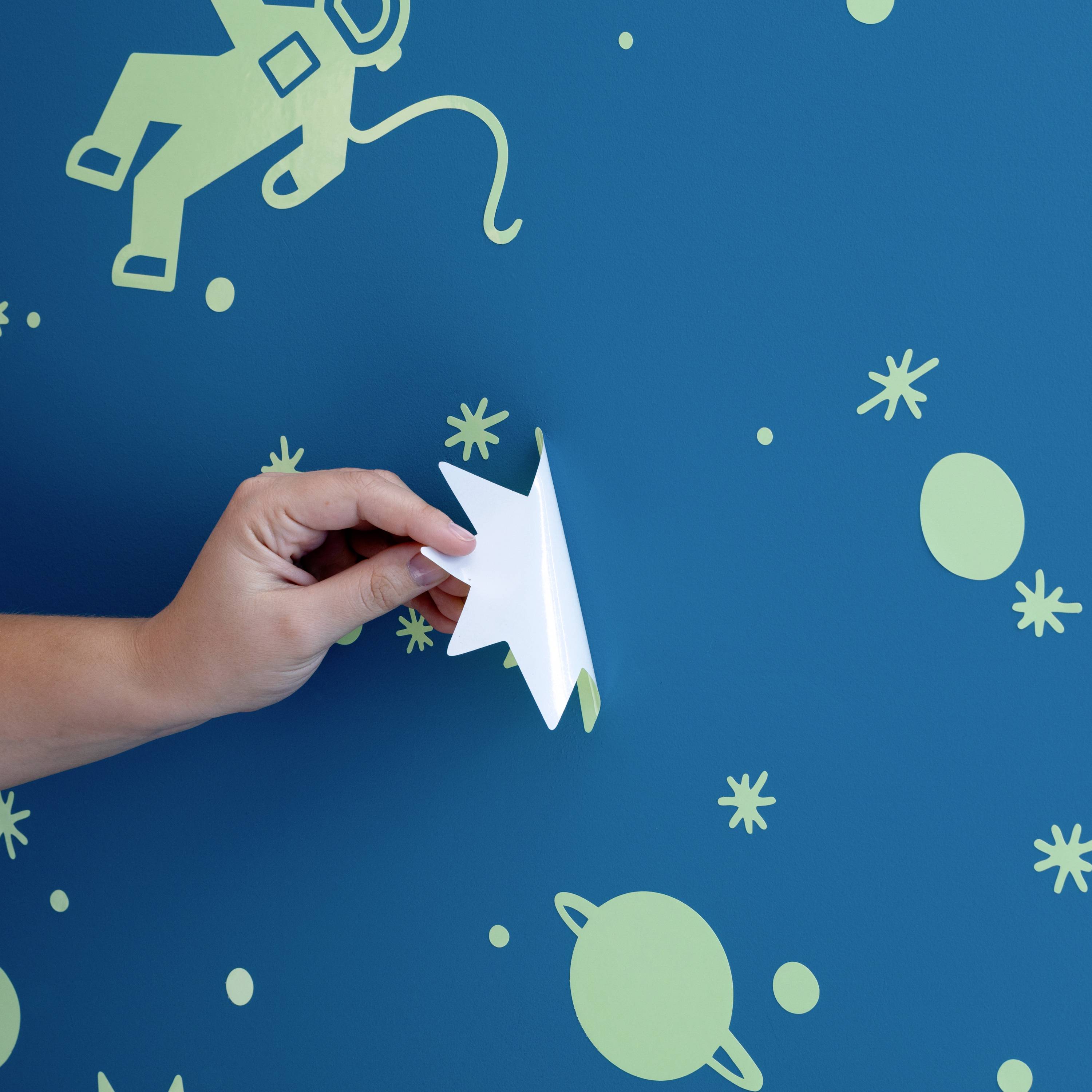 Une main tient un avion en papier blanc devant un mur bleu avec des motifs spatiaux verts tels que des astronautes et des planètes.