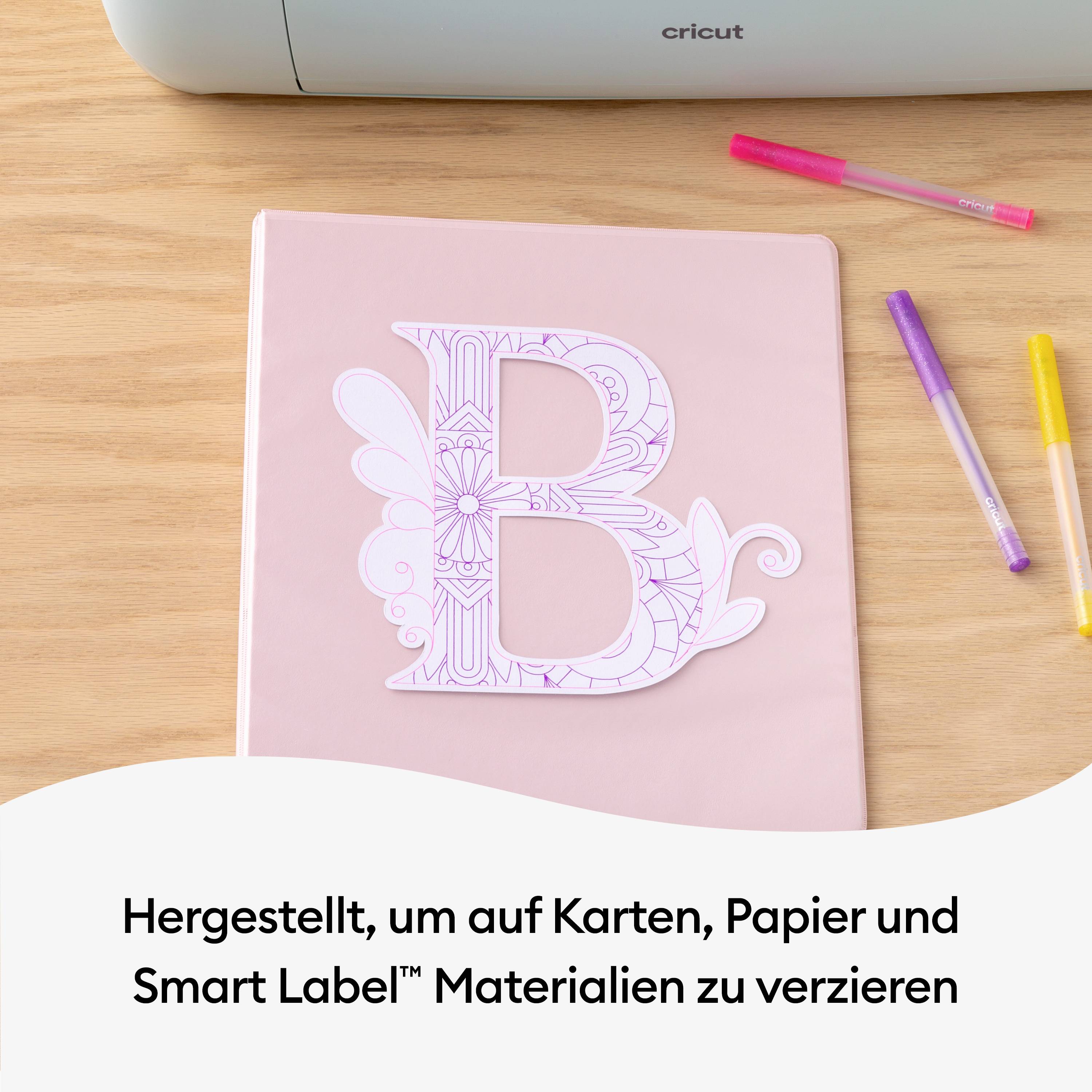 Un morceau de papier rose avec une lettre 'B' découpée, décoré d'un motif floral, est posé à côté de stylos sur une table en bois.