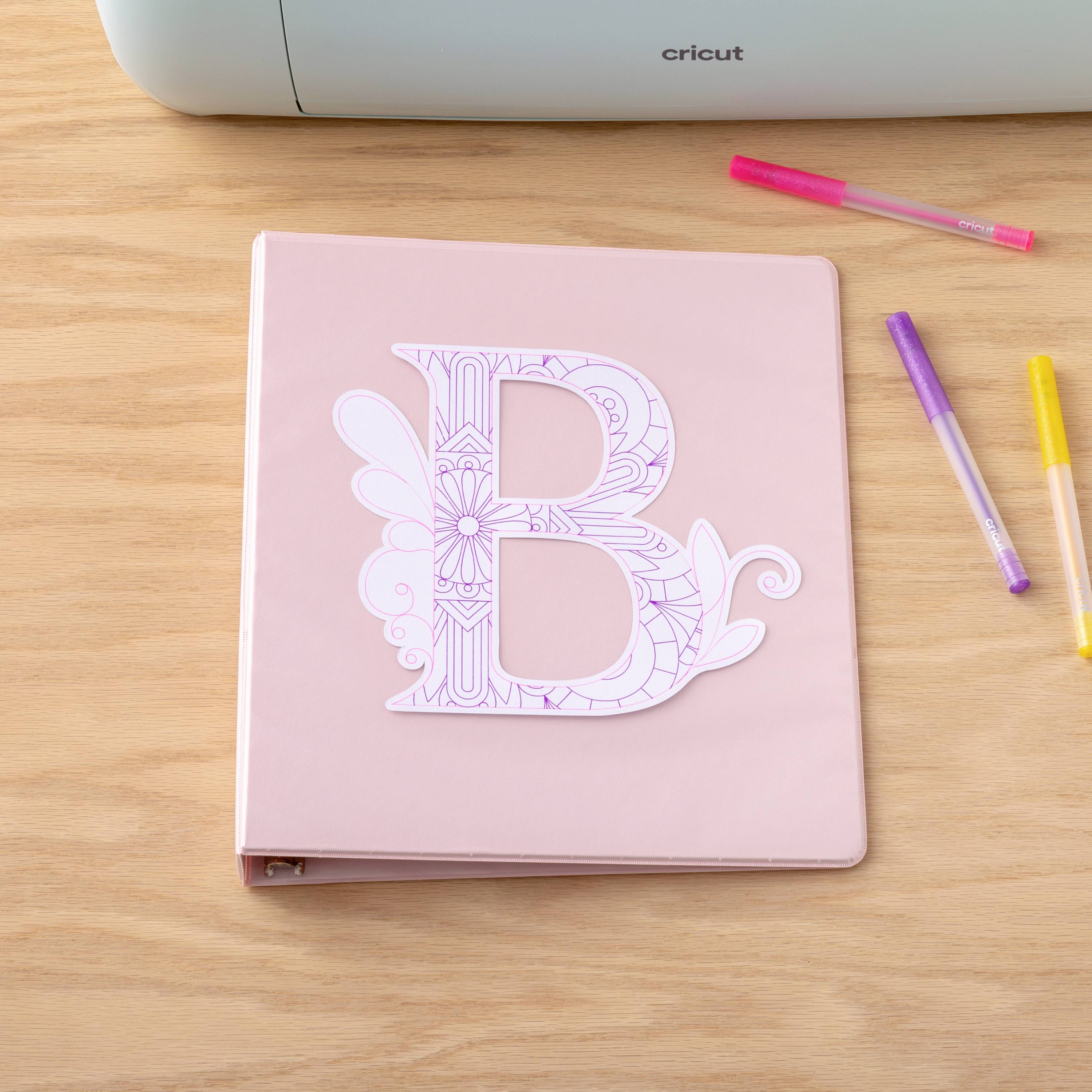 Un dossier rose avec la lettre décorative 'B' est posé sur une table en bois, entouré de stylos colorés.