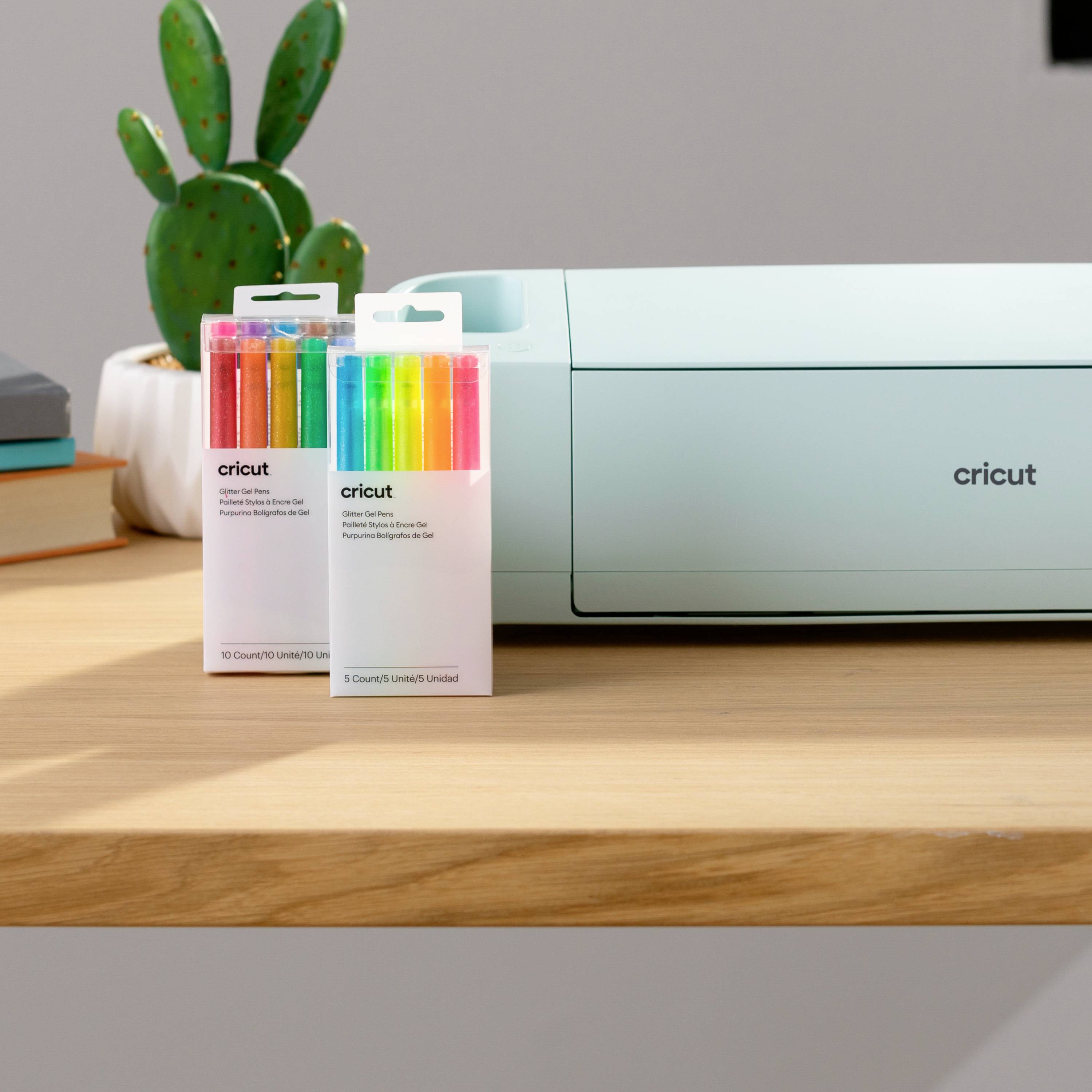 'Traceur de découpe Cricut sur une table avec deux paquets de stylos colorés à côté. Un cactus en pot est visible en arrière-plan.'