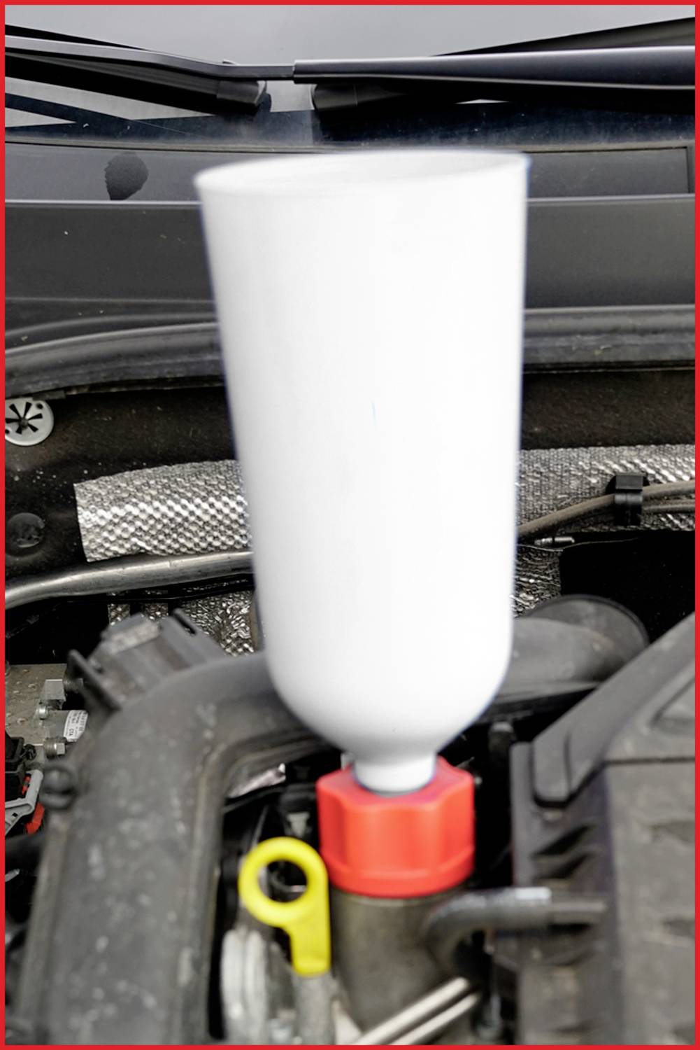 Un capot ouvert avec un entonnoir en plastique inséré dans le moteur, surmonté d'un bouchon rouge.
