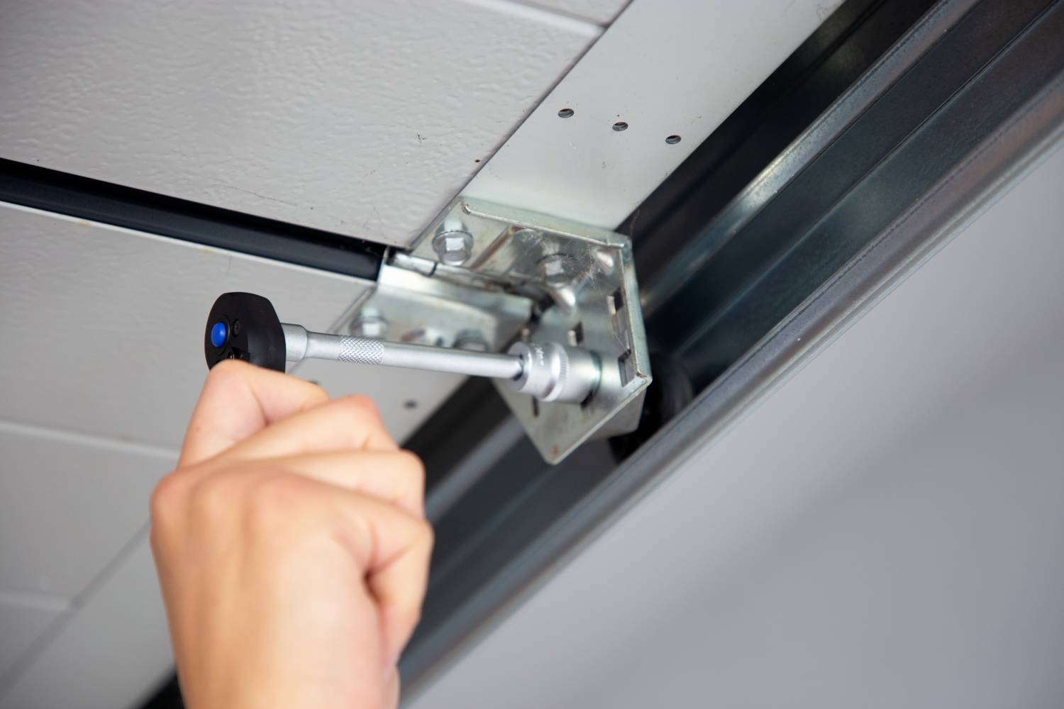 Une main utilise un outil pour réparer un garage. La personne resserre une vis sur un support métallique fixé au plafond.