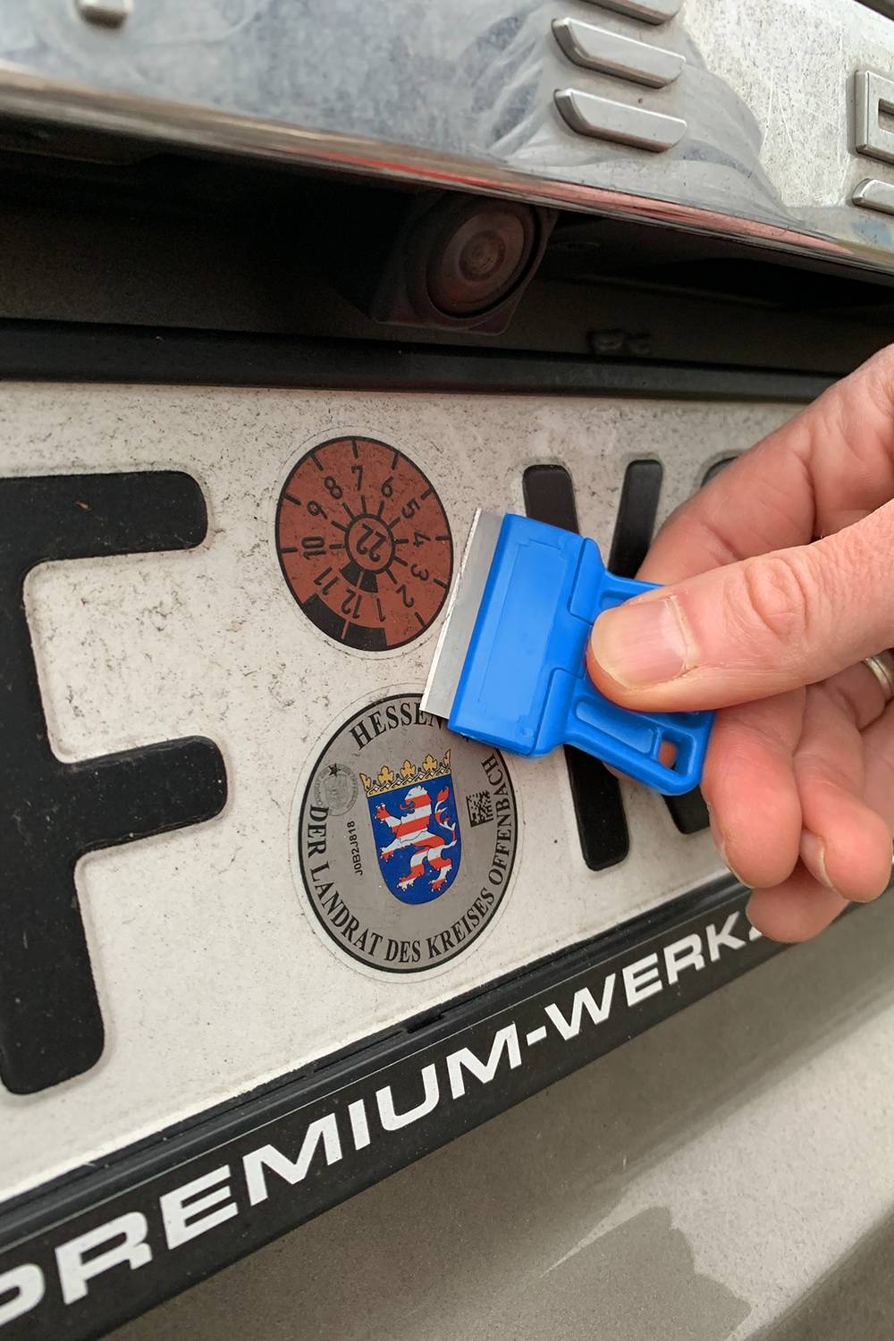 Une main retire des autocollants HU d'une plaque d'immatriculation à l'aide d'un grattoir bleu, avec en dessous les armoiries de la Hesse et le texte 'LE PRÉSIDENT DU DISTRICT'.