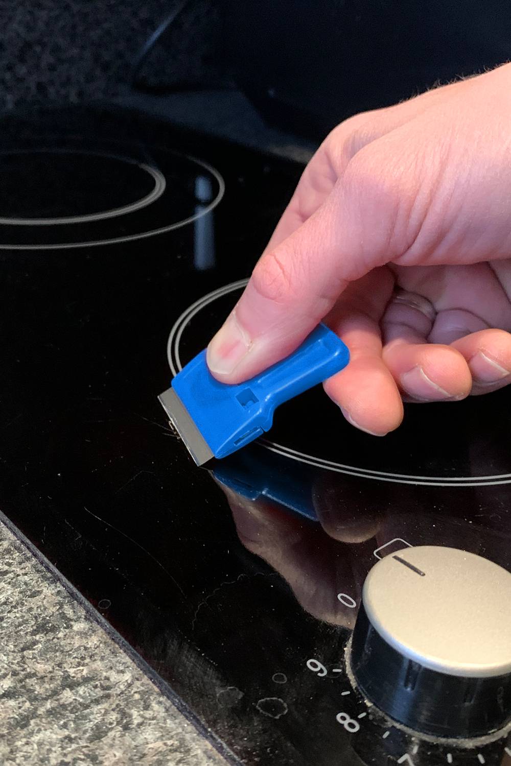 Une main tient un grattoir et nettoie une plaque de cuisson vitrocéramique. Le bouton de la plaque est réglé sur zéro.