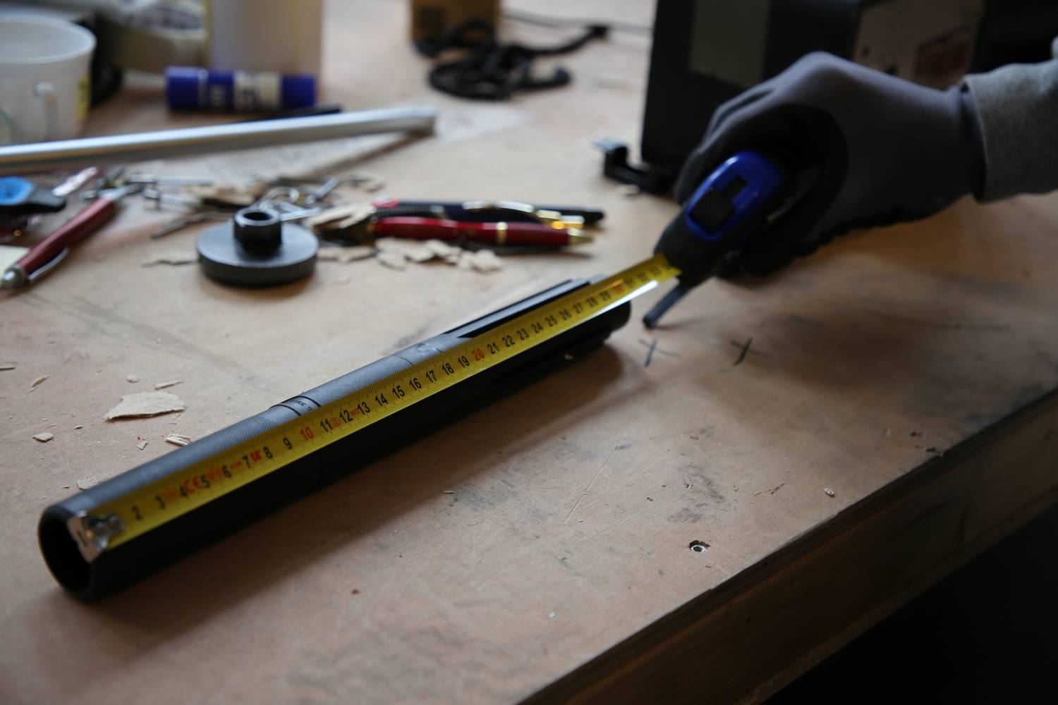 Une personne mesure un tuyau avec un mètre ruban sur une table d'atelier. À l'arrière-plan, des outils et des matériaux sont dispersés.