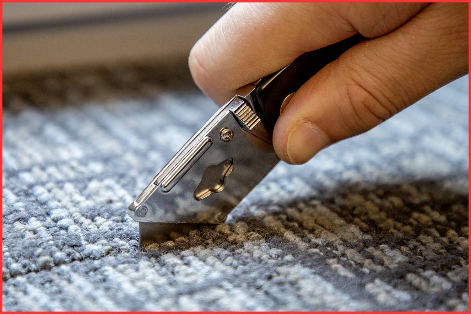 Une main coupe un tapis avec un couteau à découper. La lame du couteau est argentée, et le tapis présente un motif gris-bleu.