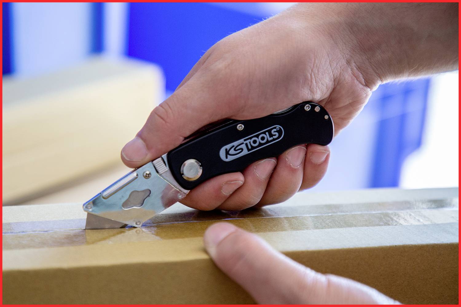 Une main tient un cutter de la marque 'KS Tools' et coupe du ruban adhésif sur un carton. L'accent est mis sur l'utilisation de l'outil.
