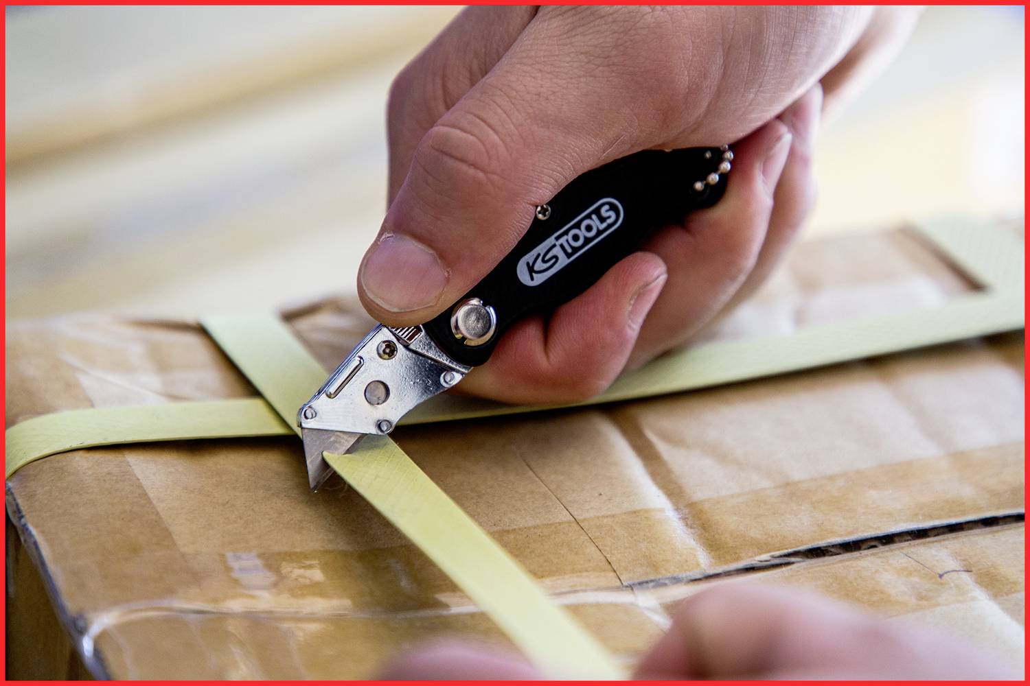 Une main coupe un élastique tendu sur un carton à l'aide d'un couteau pliant.