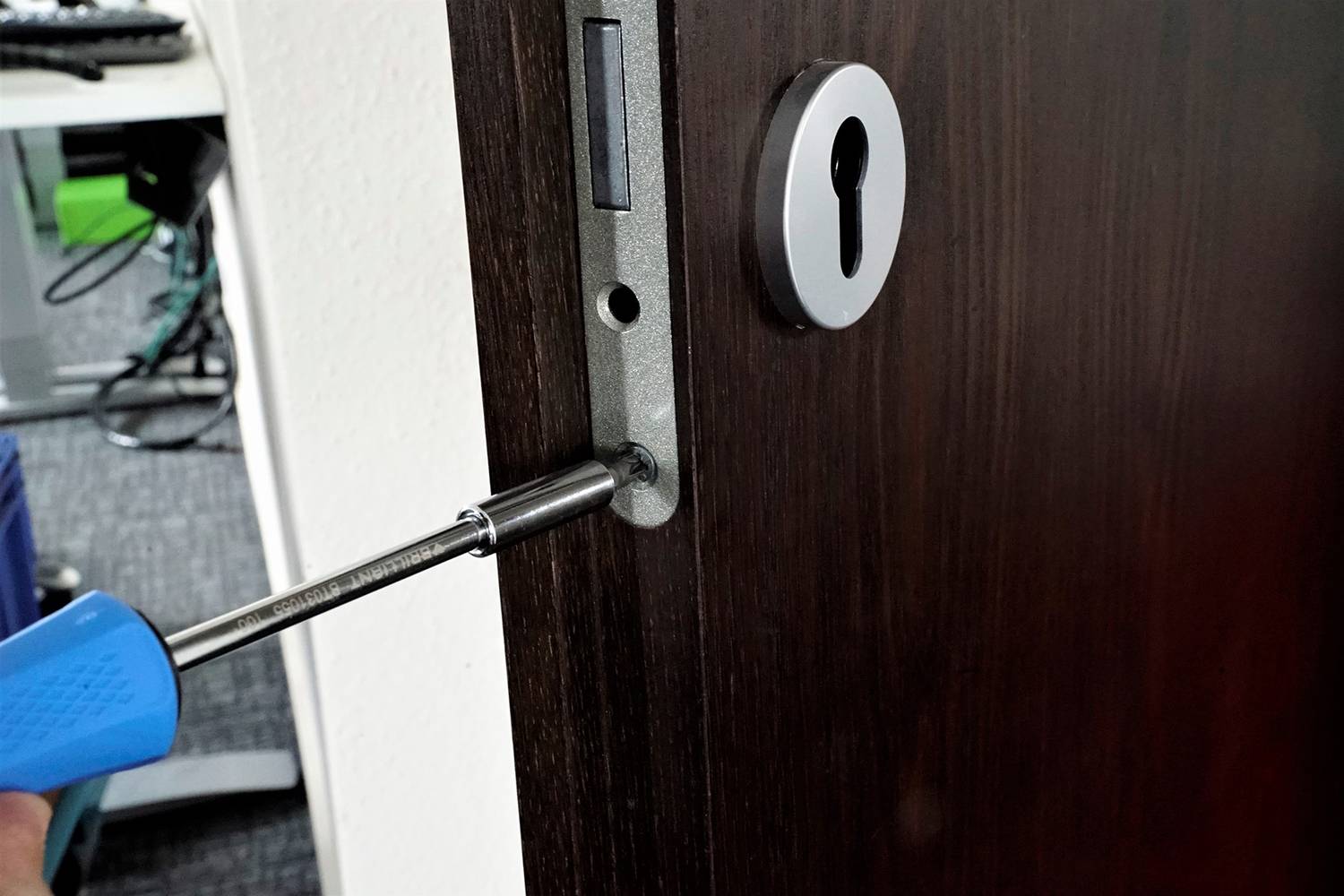 Une personne répare une serrure de porte en bois en serrant une vis avec un tournevis bleu. Un bureau est visible en arrière-plan.
