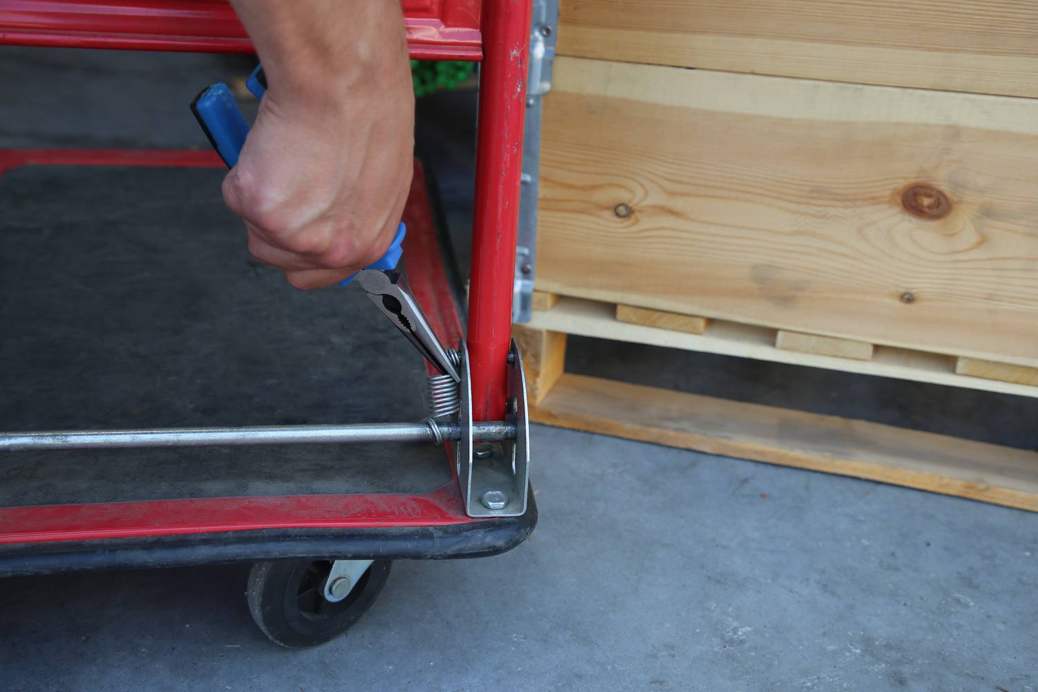 'Une main munie d'une pince répare le châssis métallique d'un chariot de transport rouge à côté d'une caisse en bois sur un sol en béton.'
