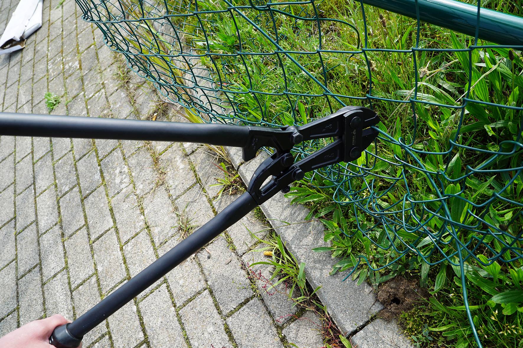 Une personne utilise un coupe-boulons pour couper une clôture grillagée verte, à côté d'une surface bétonnée.
