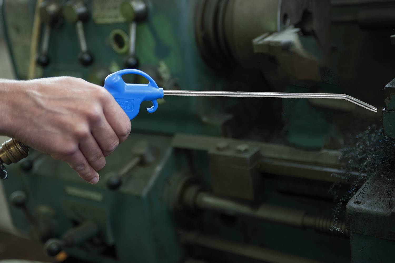 Une main tient un pistolet à air comprimé bleu, dirigé vers une machine industrielle pour la nettoyer ou la refroidir.