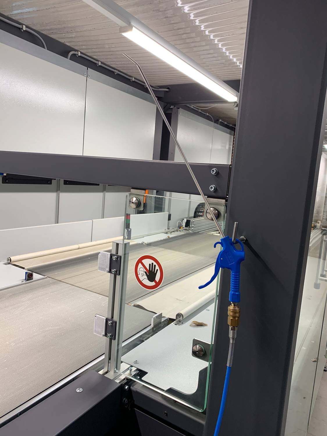 Une machine dans un hall de production avec un couvercle en verre et un panneau d'avertissement 'Éloigner les mains'. Un pistolet à air comprimé bleu est suspendu sur le côté.