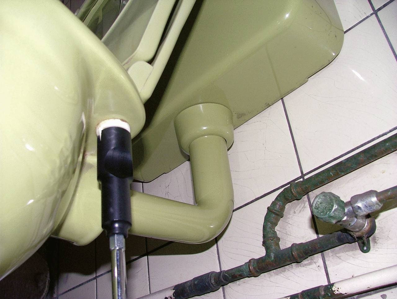 Lavabo vert vu de dessous, montrant le tuyau de vidange et les canalisations adjacentes sur un fond de carrelage.