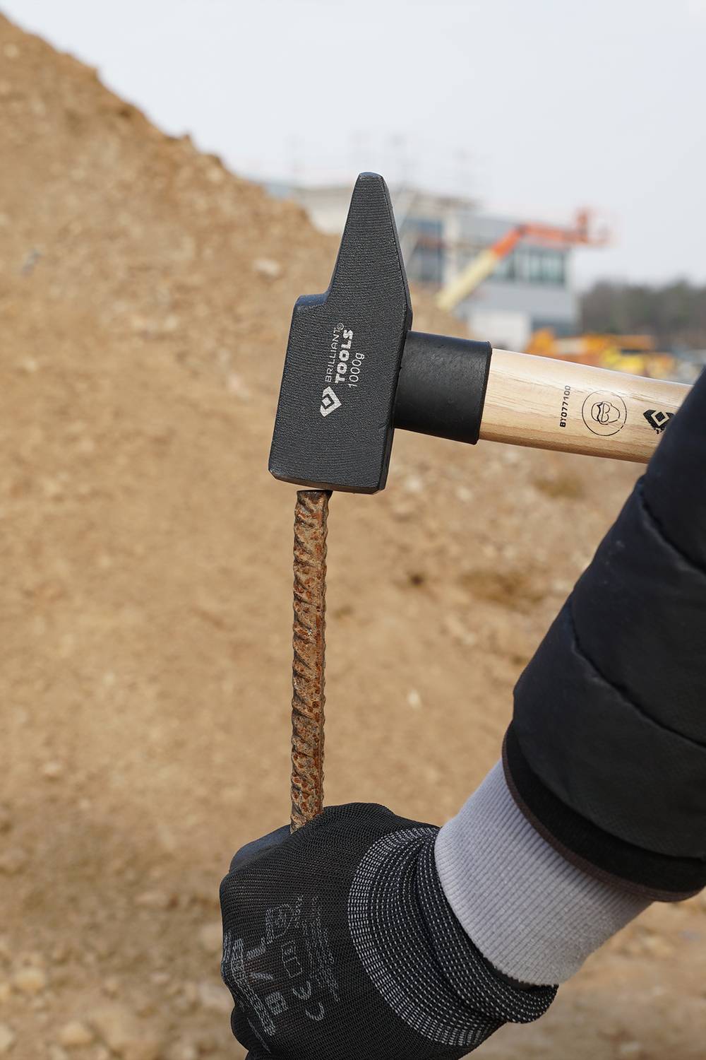 Une main gantée tient un marteau frappant sur une tige de chantier. En arrière-plan, des tas de terre et des bâtiments flous sont visibles.