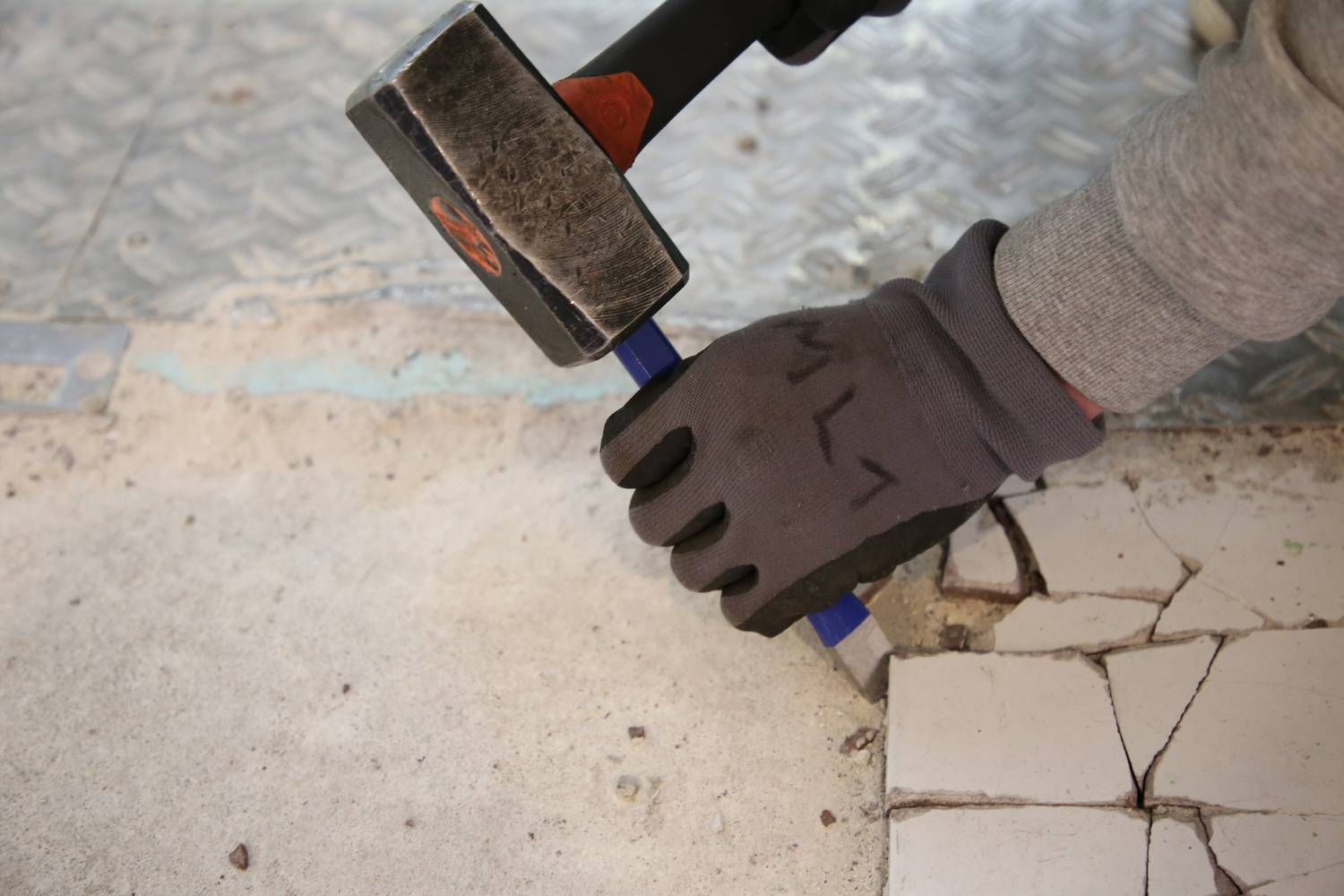 Une personne retire des carreaux d'un sol à l'aide d'un marteau et d'un burin. Elle porte des gants et travaille sur une surface rugueuse.