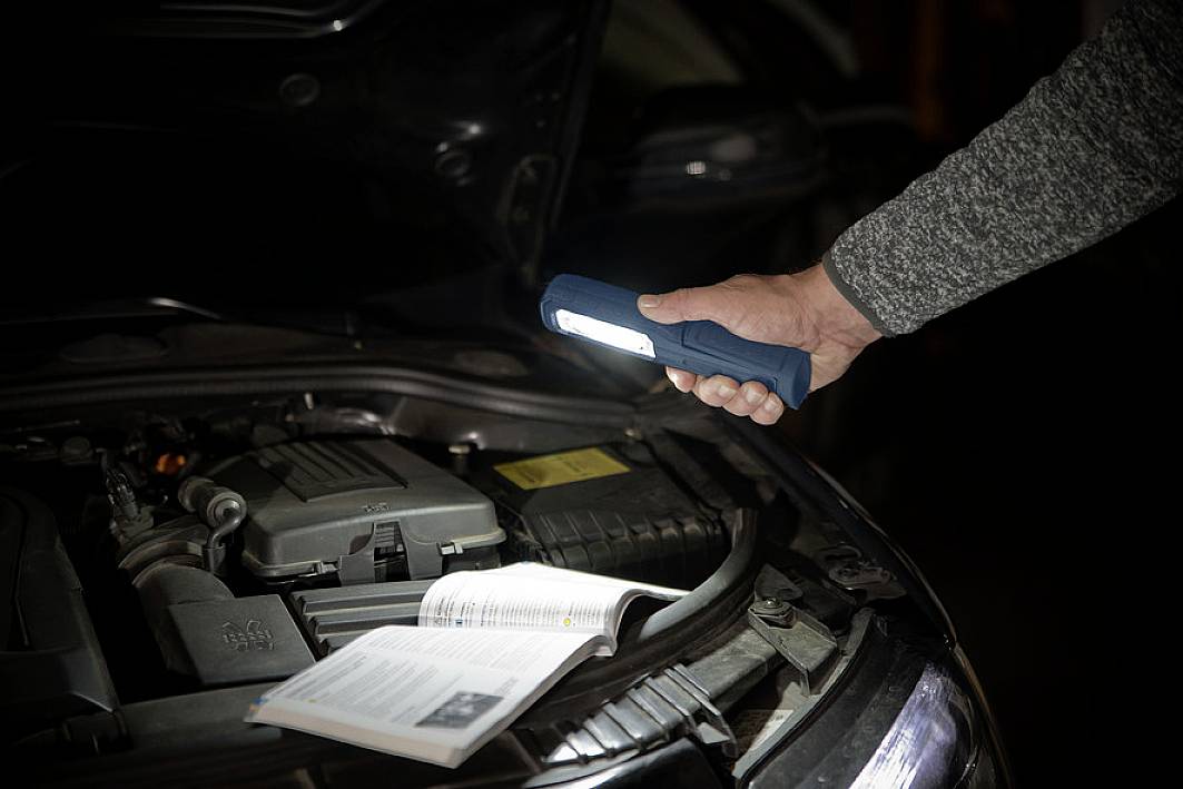 Une personne tient une lampe de poche au-dessus du capot ouvert d'une voiture, éclairant le moteur et le manuel.