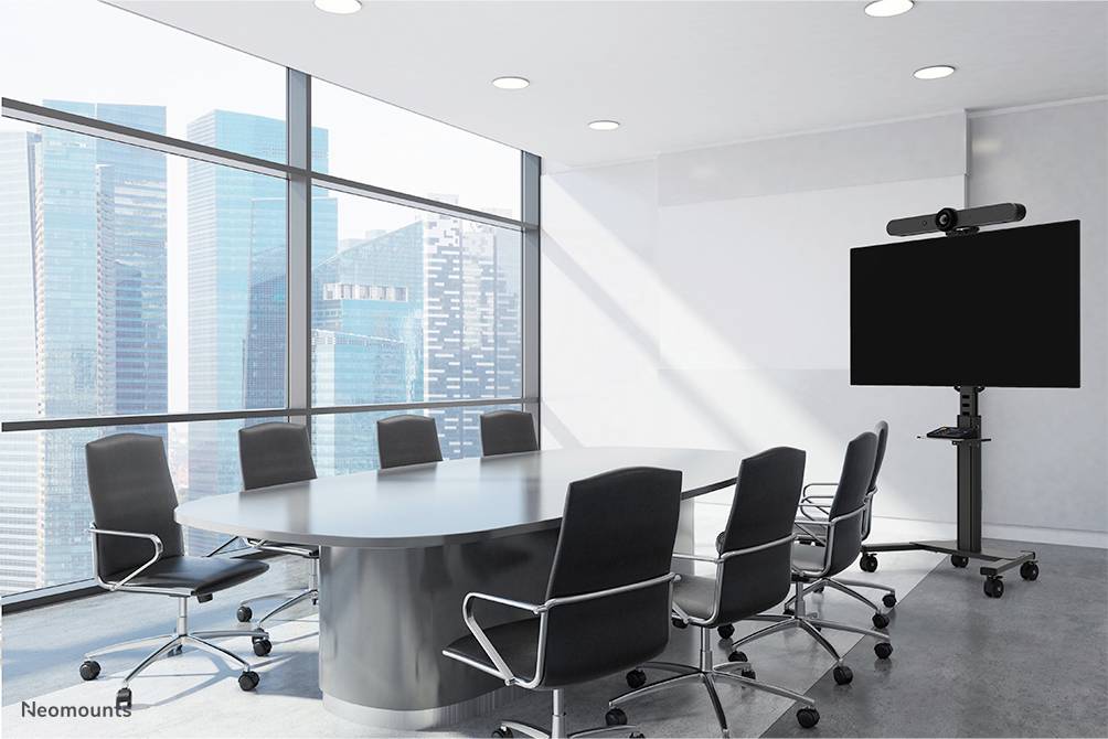 Salle de conférence avec une grande table ovale, huit chaises noires et un grand écran sur support ; fenêtre offrant une vue sur la ville en arrière-plan.