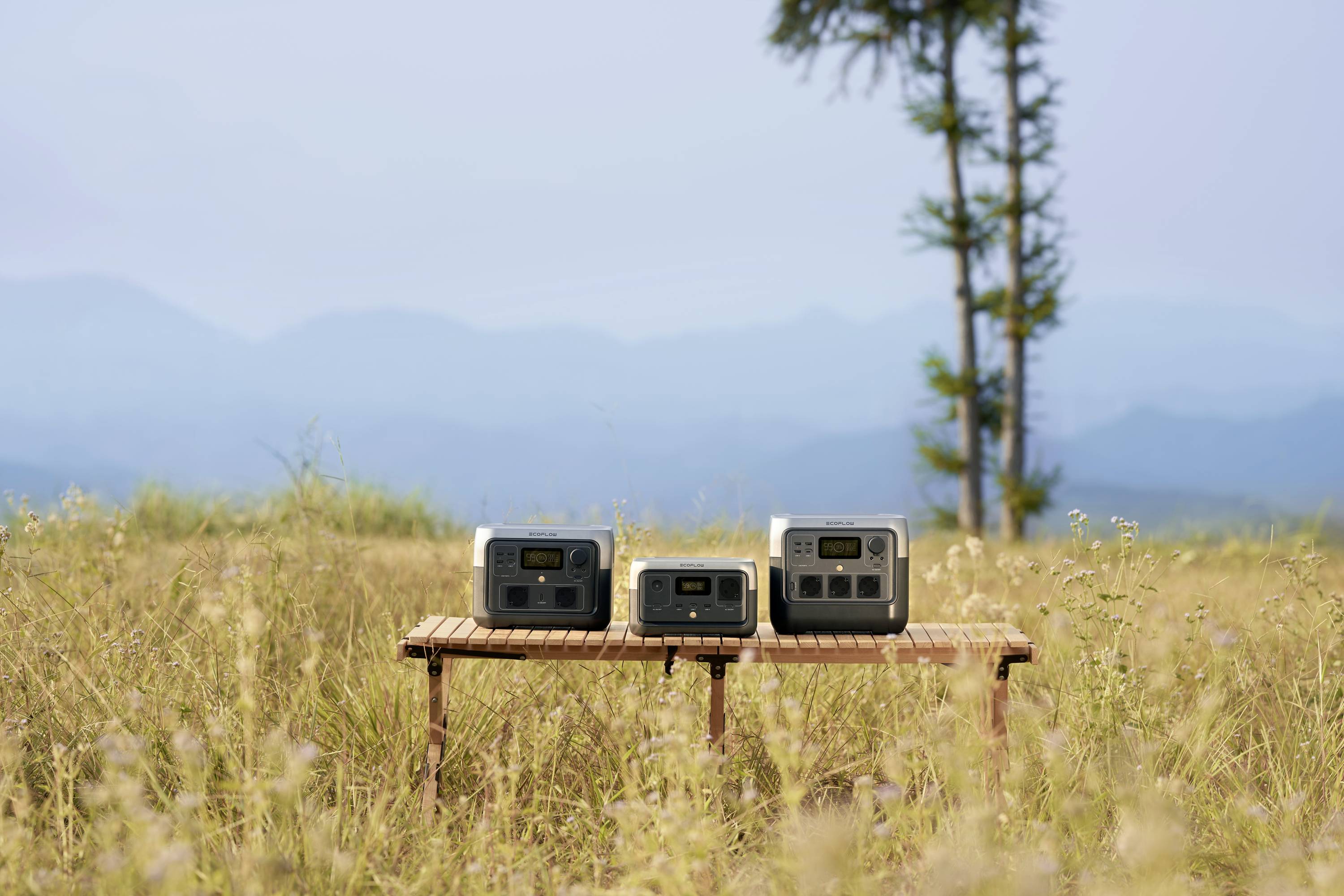 Trois stations d'énergie portables sont posées sur une table en bois dans une prairie, avec un arbre et des montagnes en arrière-plan.