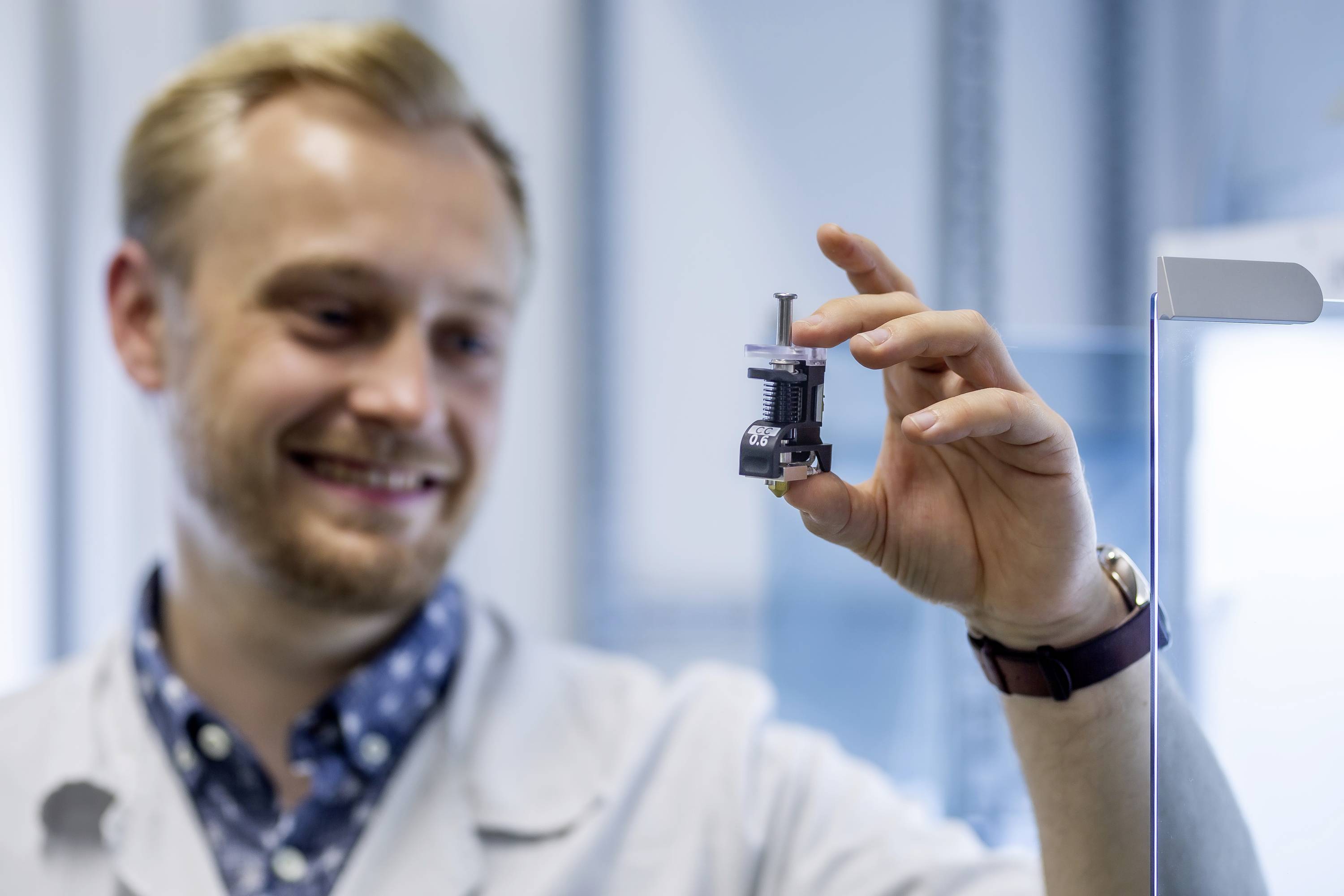 Un homme dans un laboratoire tient un petit dispositif technique et sourit. L'arrière-plan est flou. La focus est sur l'appareil qui est soigneusement inspecté.