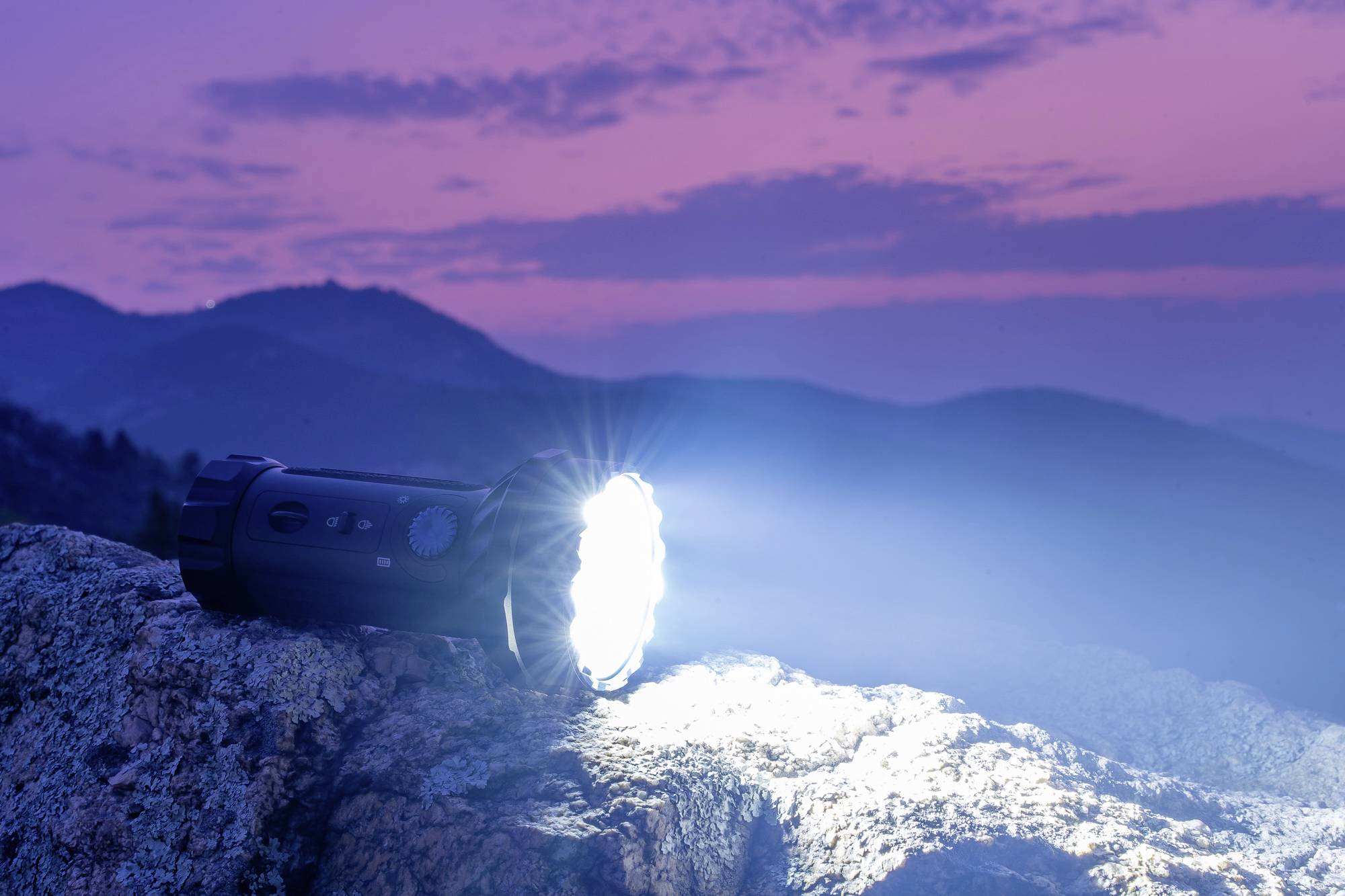 Une lampe de poche brille sur un rocher au crépuscule, entourée de collines sous un ciel lilas et rose.