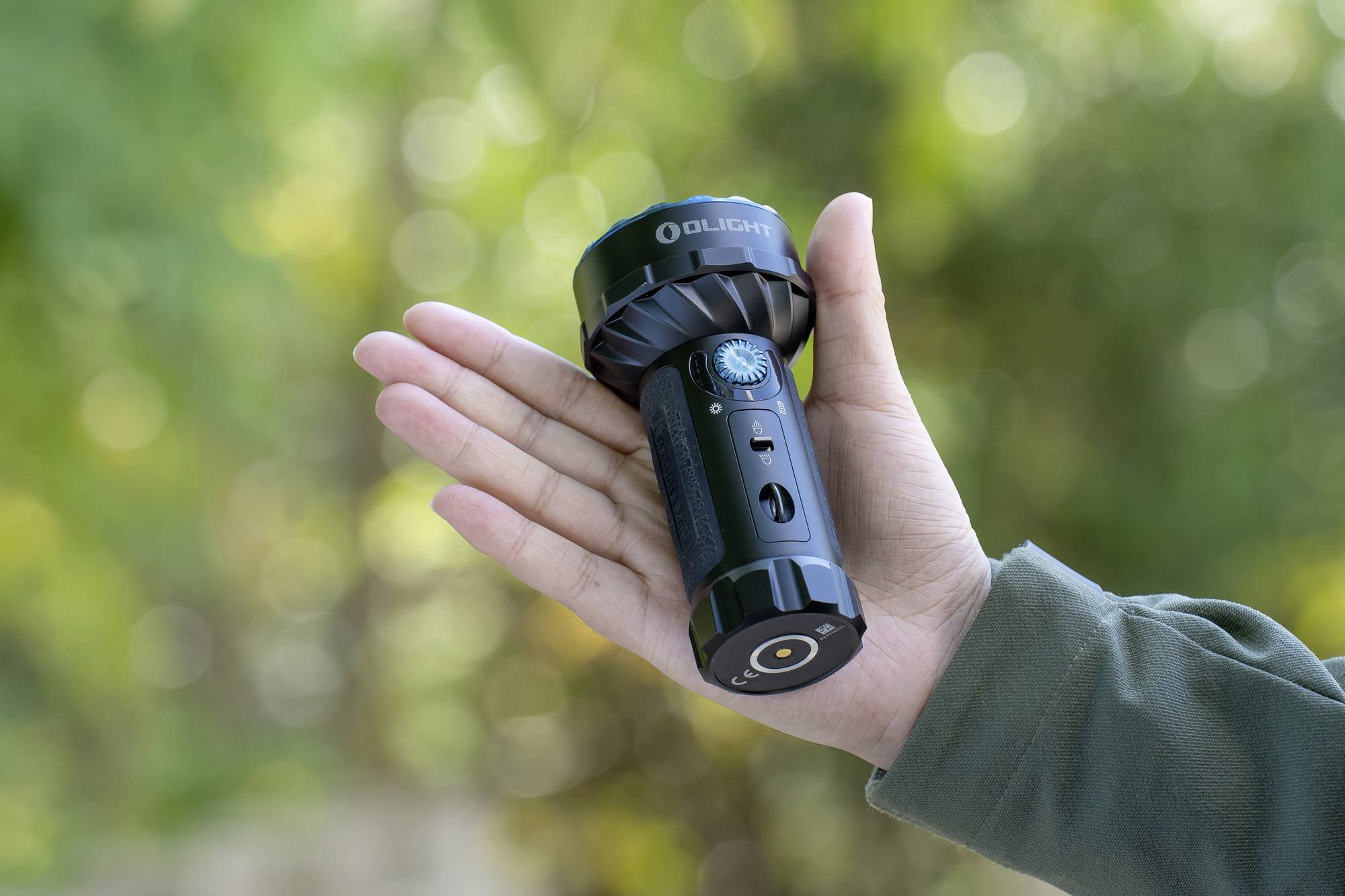 Une main tient une lampe de poche noire avec un bouton bleu, un arrière-plan vert flou en profondeur.
