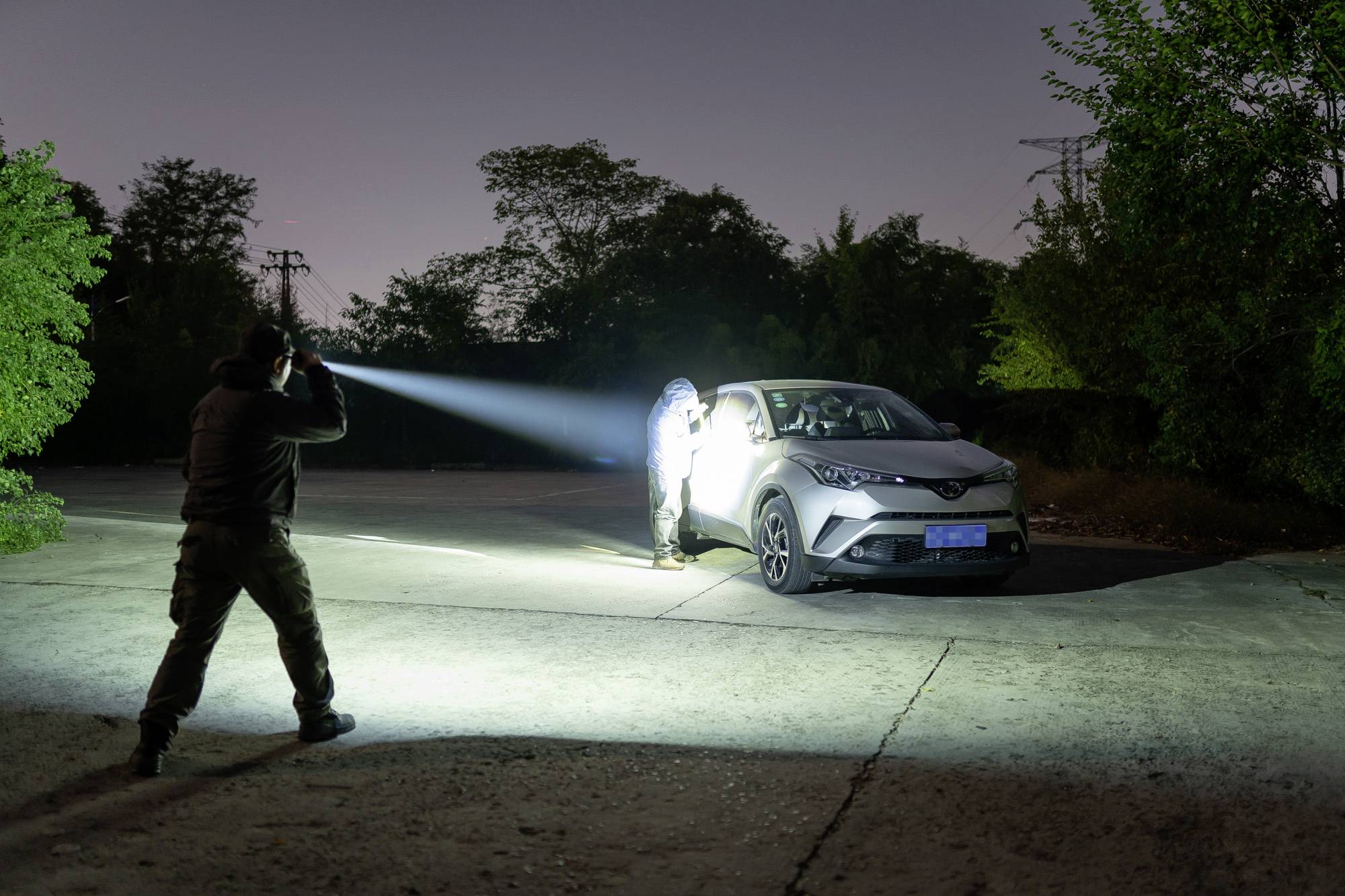 'Deux personnes inspectent une voiture la nuit à l'aide de lampes de poche sur un parking désert, entouré d'arbres.'