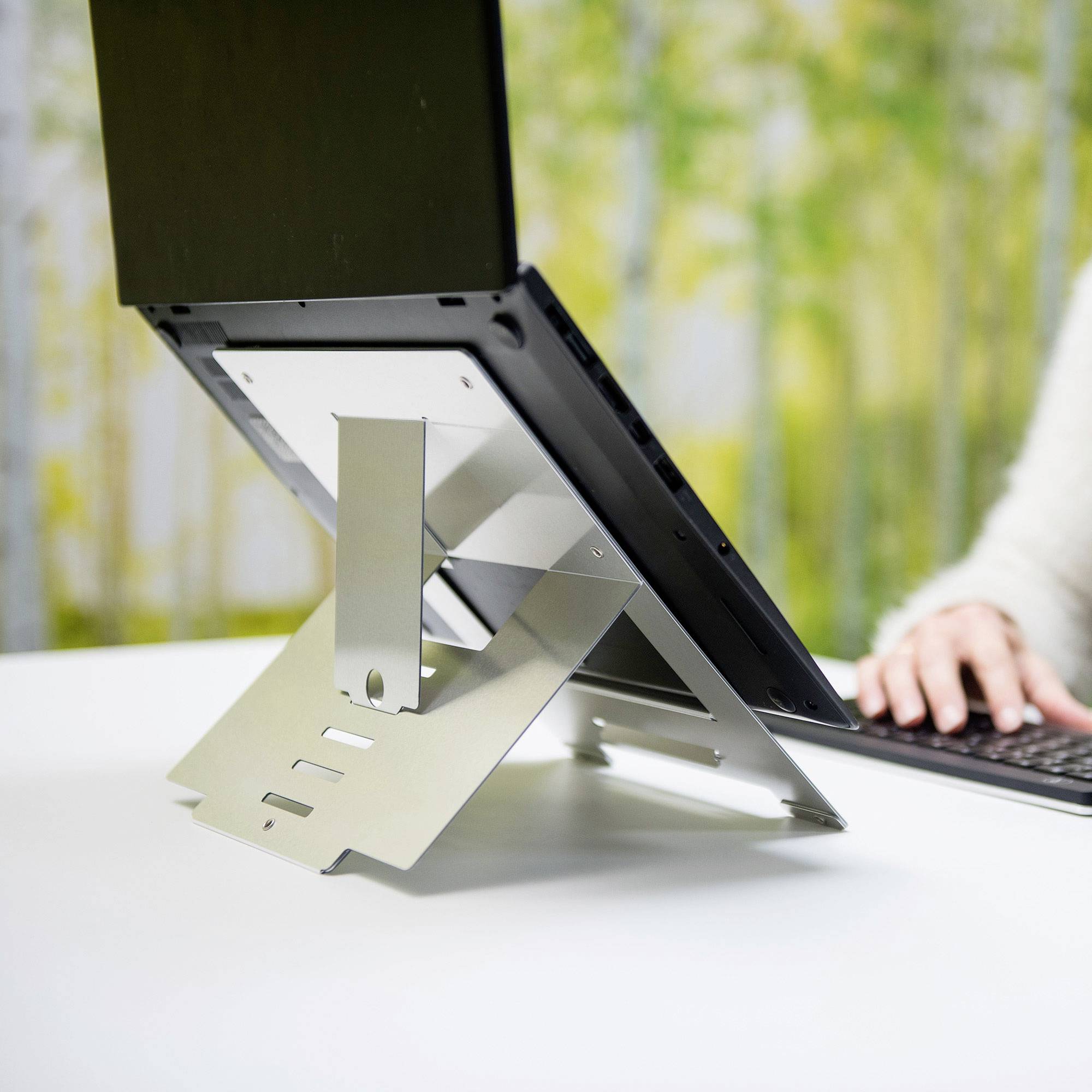 Un ordinateur portable est posé sur une table, installé sur un support métallique réglable pour augmenter la hauteur de l'écran, dans une pièce lumineuse avec un arrière-plan naturel.