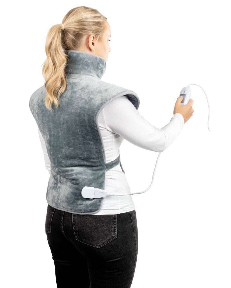 Une femme porte un gilet chauffant électrique sur le dos et tient la télécommande à la main. Le gilet est gris et couvre la nuque et les épaules.