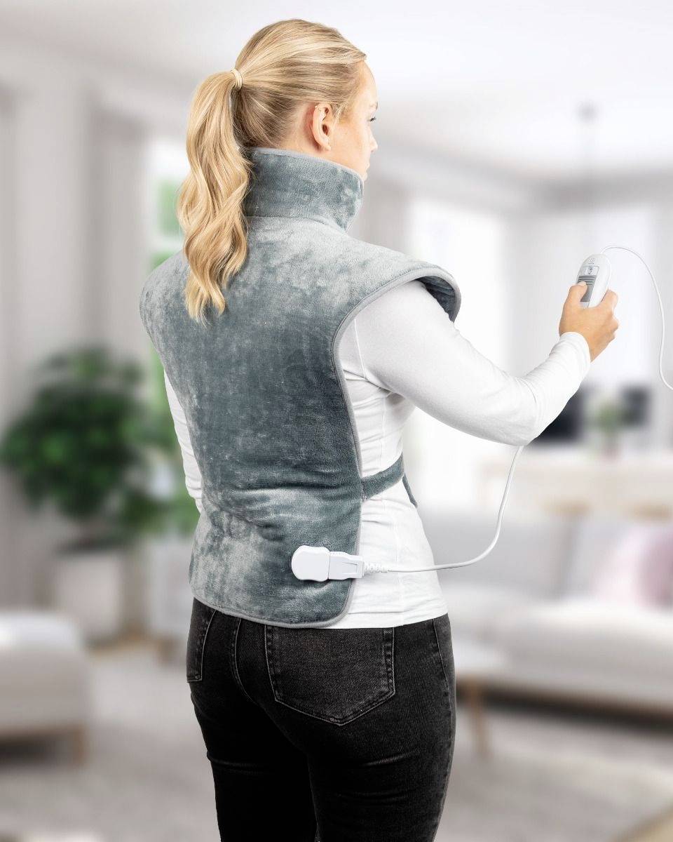 Une femme se tient dans un salon et porte un coussin chauffant électrique sur les épaules et le dos. Elle tient la télécommande du coussin chauffant.