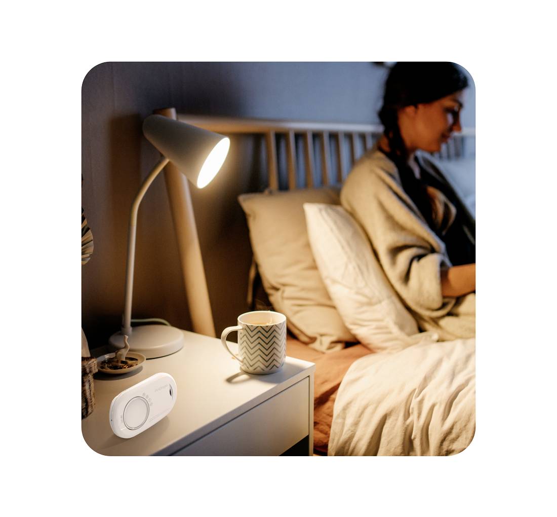 Une femme est assise dans son lit avec un ordinateur portable en arrière-plan ; au premier plan, une lampe s'illumine à côté d'un interrupteur intelligent sur la table de chevet.