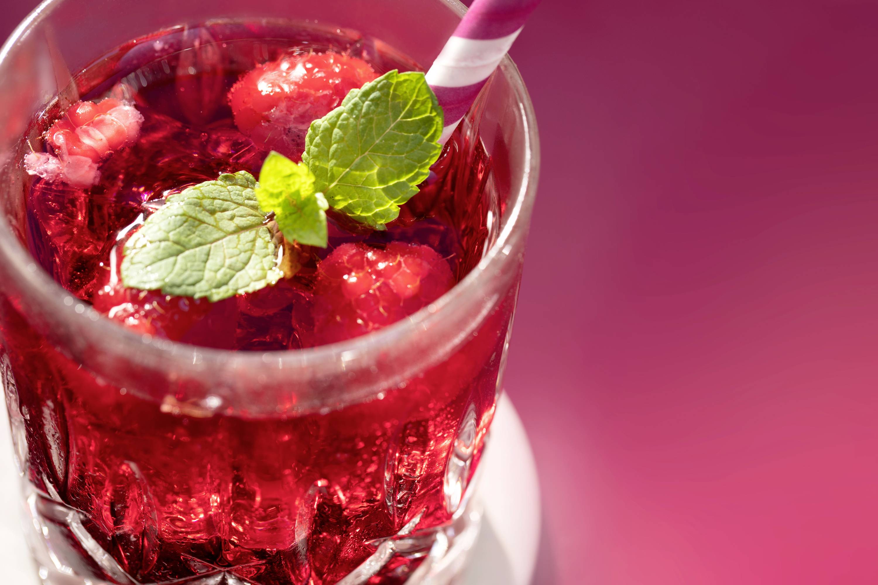 Un verre de jus de fruits rouge avec des glaçons, des feuilles de menthe et une paille rayée sur un fond rose.