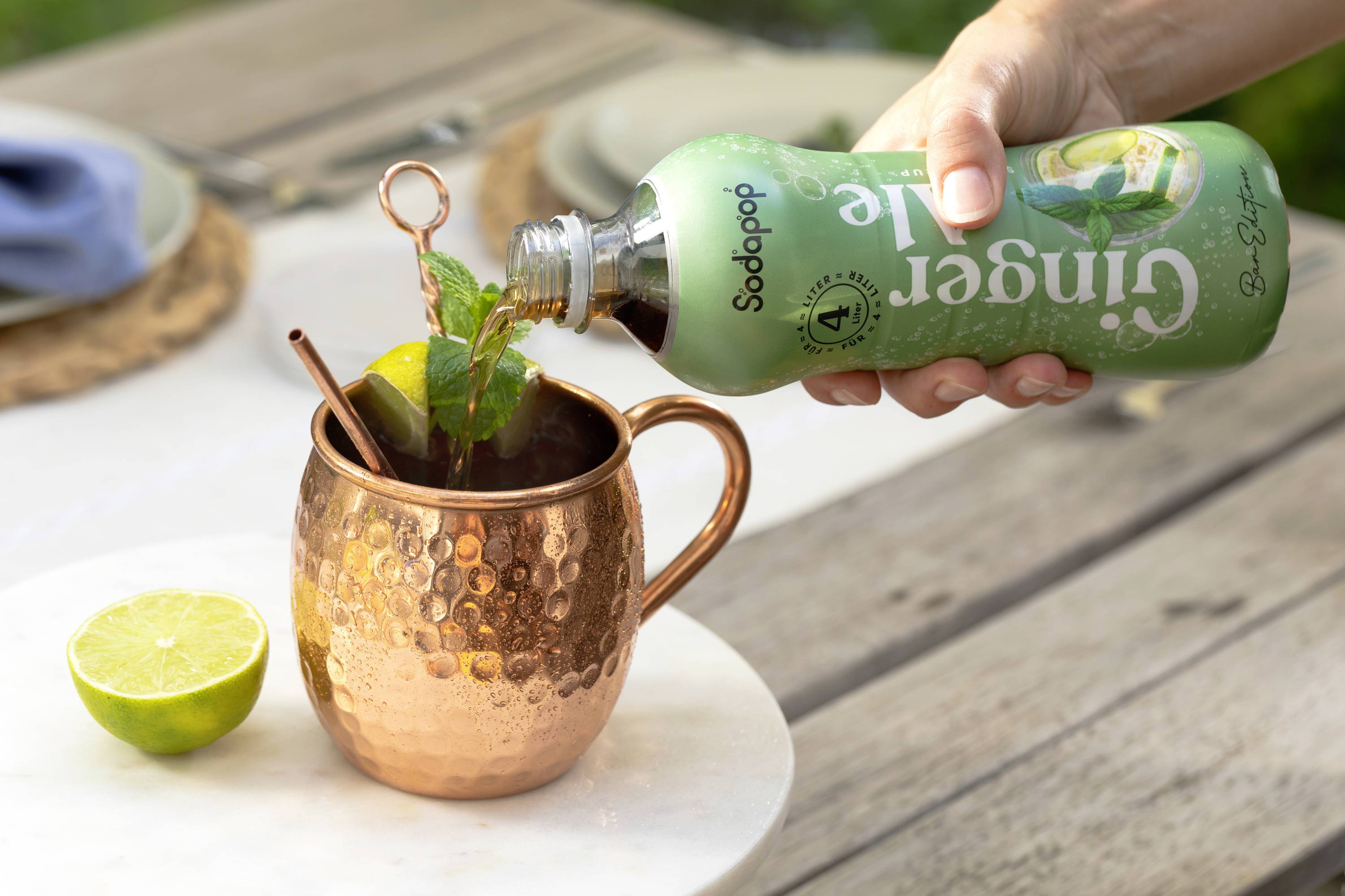 Une main verse du ginger beer d'une bouteille verte dans un gobelet couleur cuivre rempli de glaçons, de menthe et de citron posé sur une table.