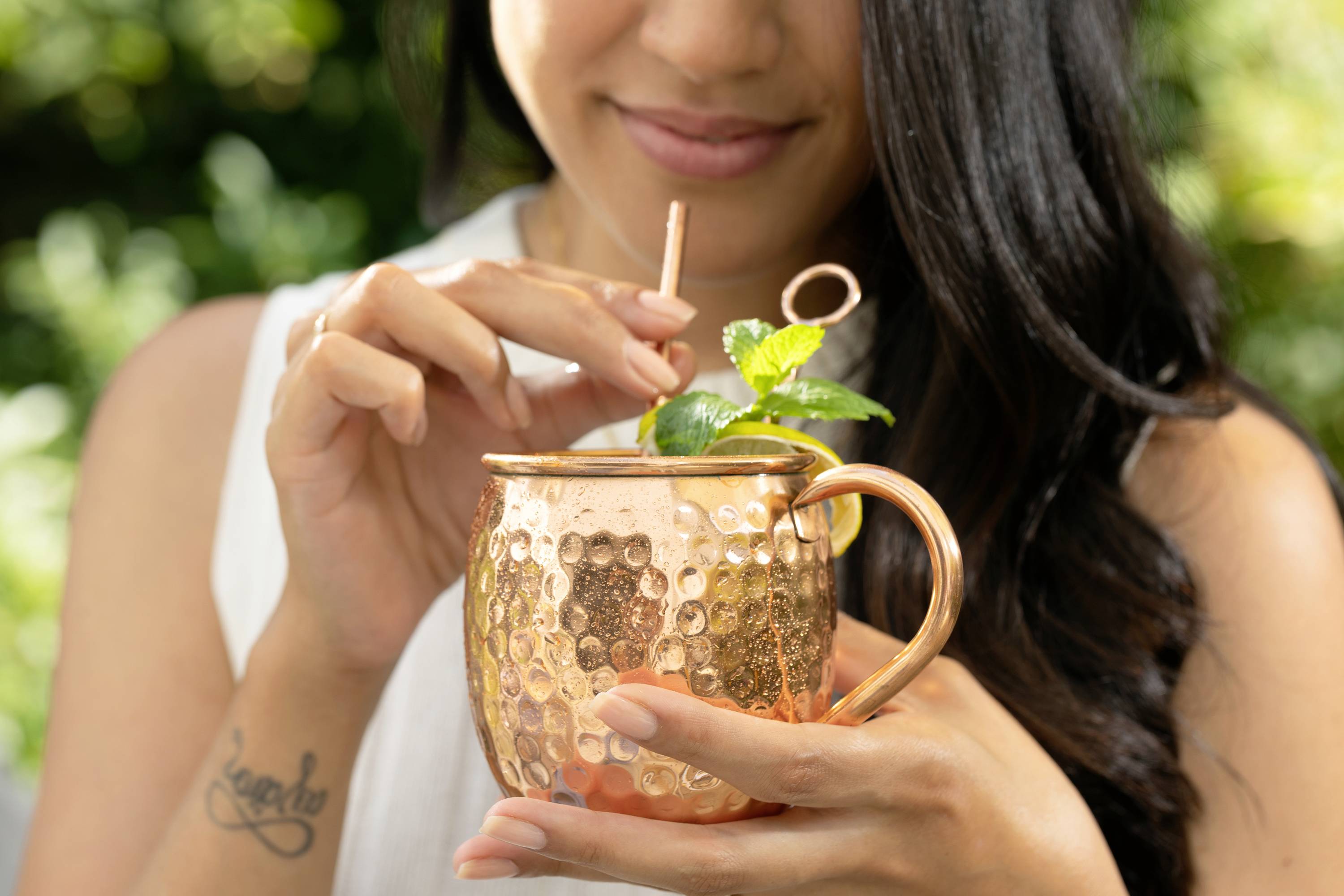 Une femme tient une tasse en cuivre contenant une boisson et des feuilles de menthe. Elle remue avec une paille.