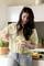 Une femme aux cheveux longs verse de l'eau d'une carafe dans un verre contenant une tranche de citron. Une cuisine est visible en arrière-plan.