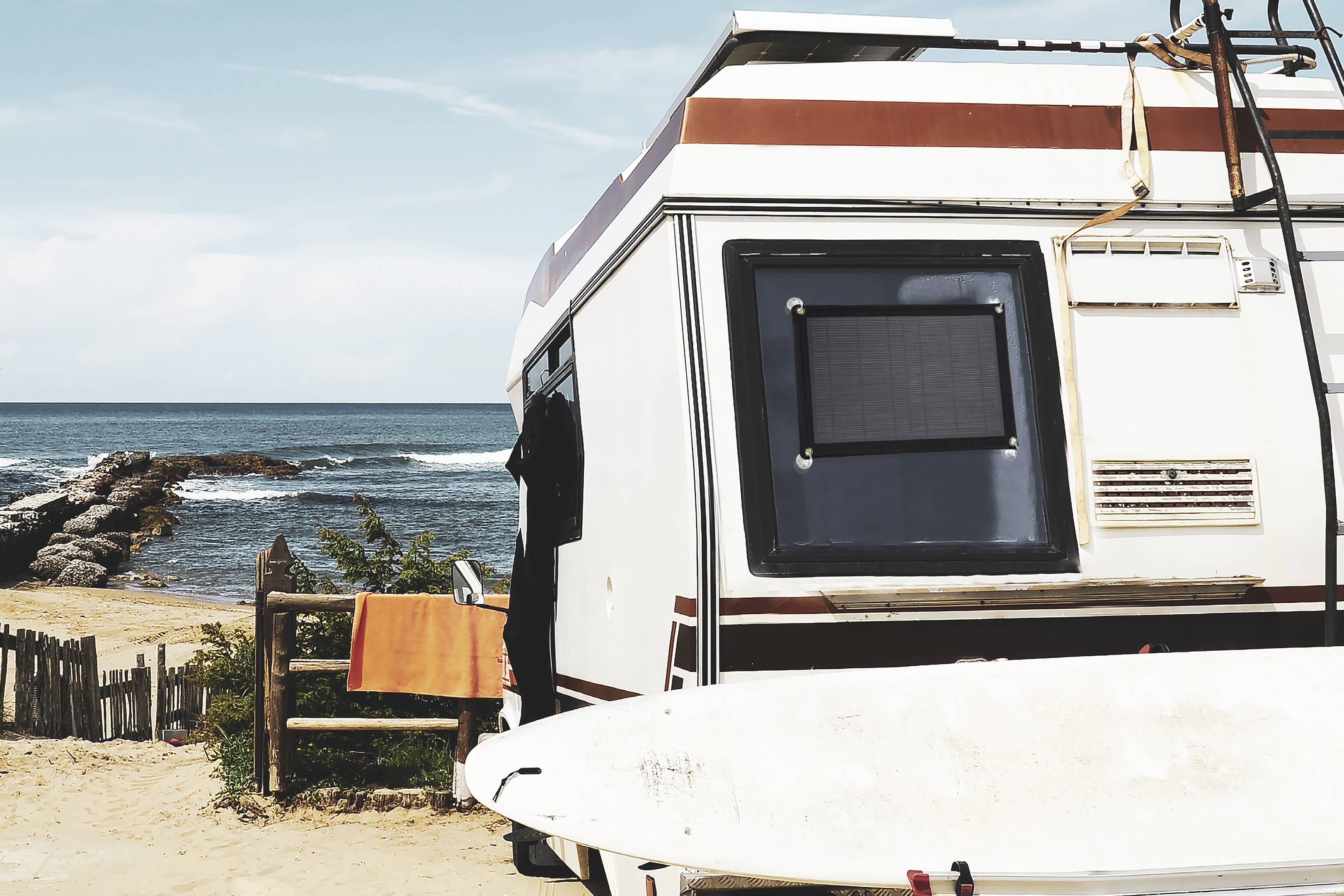 Un camping-car est stationné sur la plage avec vue sur la mer. Une planche de surf est appuyée contre le côté du véhicule, et une serviette orange est suspendue à côté de la porte.