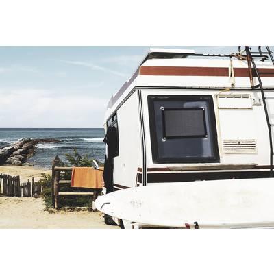 Un camping-car est stationné sur la plage avec vue sur la mer. Une planche de surf est appuyée contre le côté du véhicule, et une serviette orange est suspendue à côté de la porte.