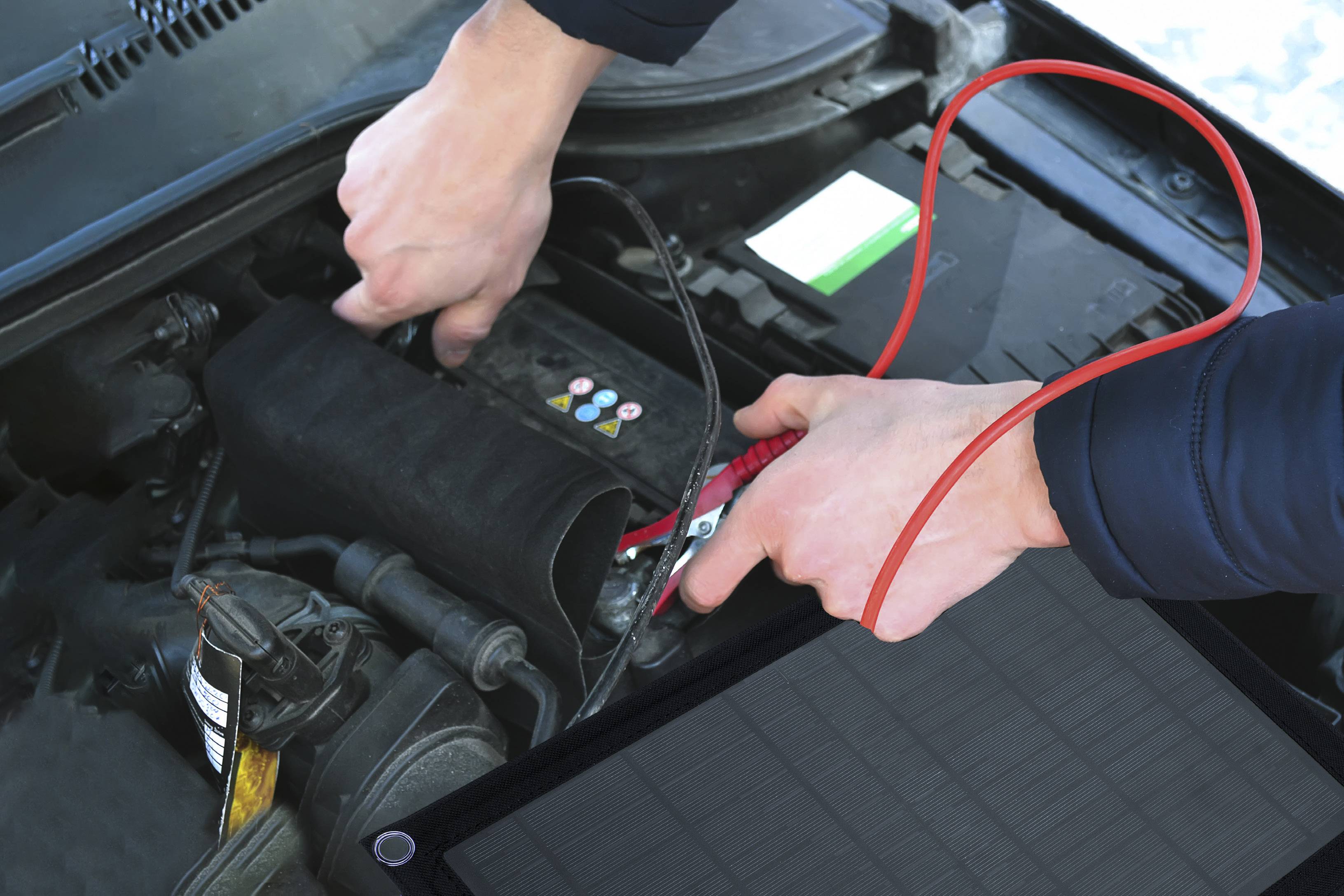 Une personne répare une batterie de voiture à l'aide de câbles de démarrage. Les mains connectent le câble rouge à la borne de démarrage du véhicule.