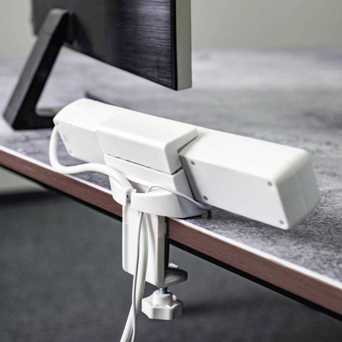 Un dispositif de gestion de câbles blanc est fixé à un bureau, avec un écran visible en arrière-plan.