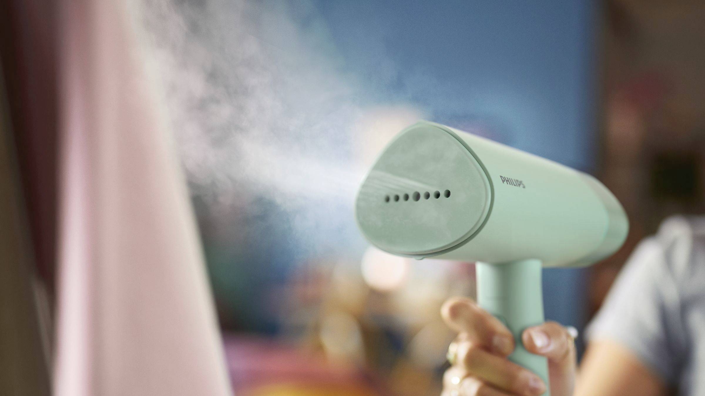 Une main tient un appareil de repassage à vapeur bleu qui vaporise de la vapeur sur une robe rose. L'arrière-plan est flou et montre un environnement de buanderie.