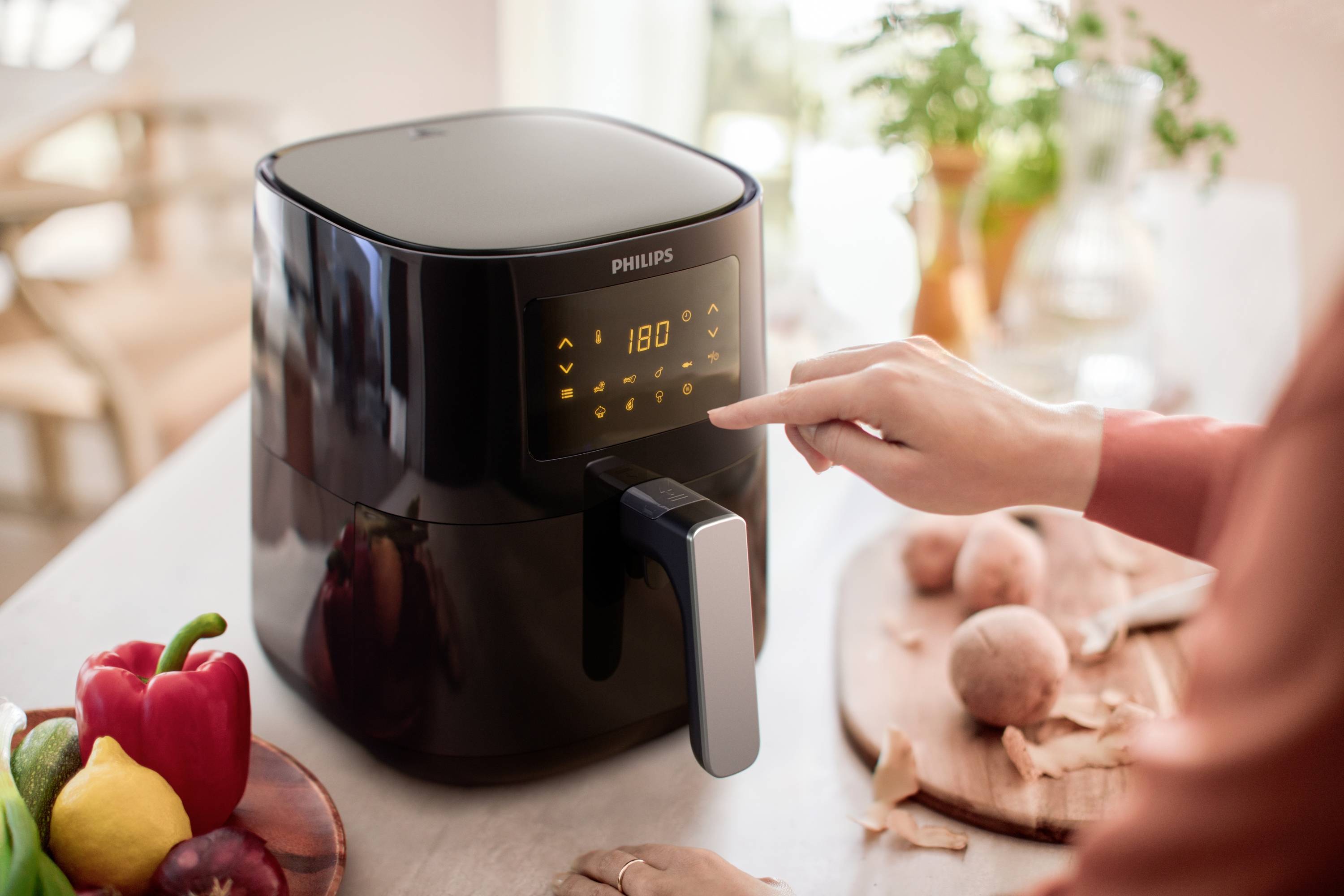 Une personne règle la température à 180 degrés sur l'affichage numérique d'une friteuse à air, entourée de légumes dans une cuisine.