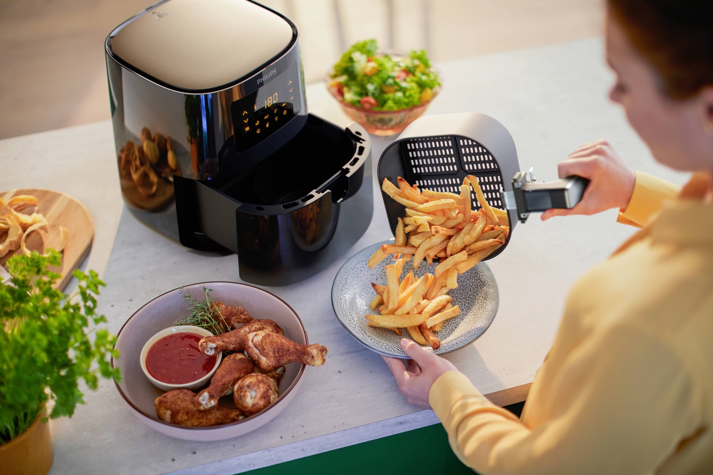 Une personne sert des frites à partir d'une friteuse à air chaud sur une assiette. Sur la table se trouvent également des cuisses de poulet et une salade.