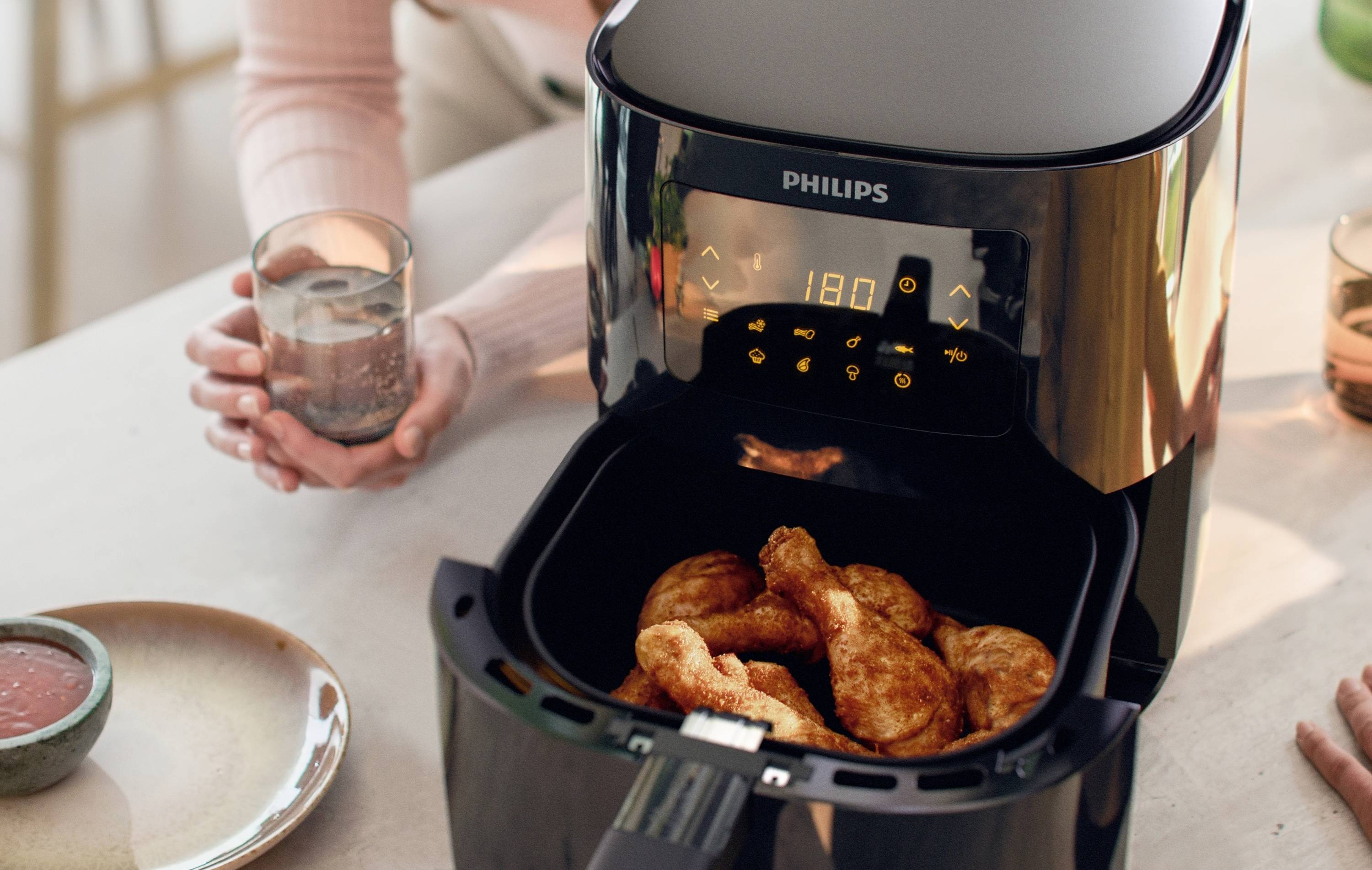 Une friteuse à air chaud posée sur une table avec des pilons de poulet dorés dans le panier de cuisson. Une personne tient un verre d'eau à côté.