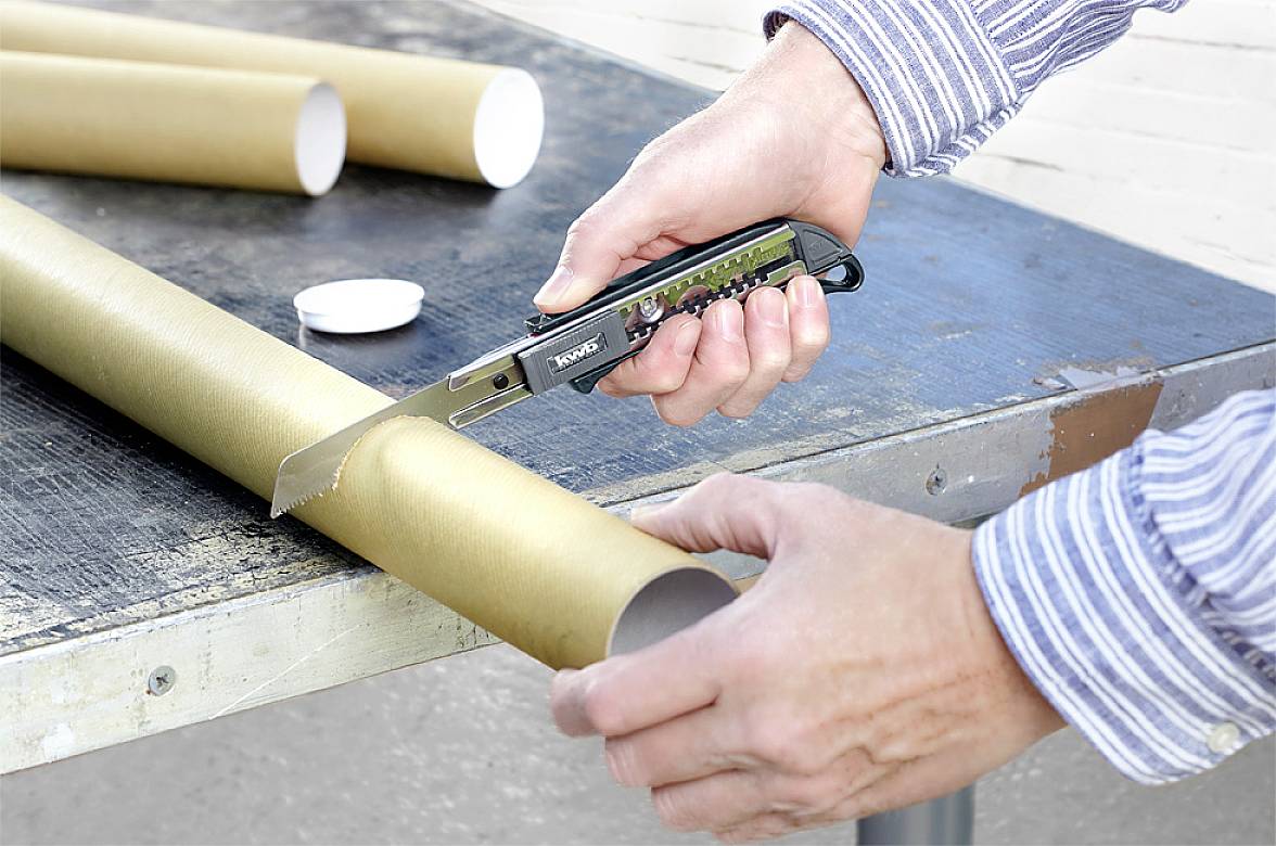 Une personne coupe un tube en carton brun avec un cutter sur une table métallique. Les mains et le couteau sont visibles.