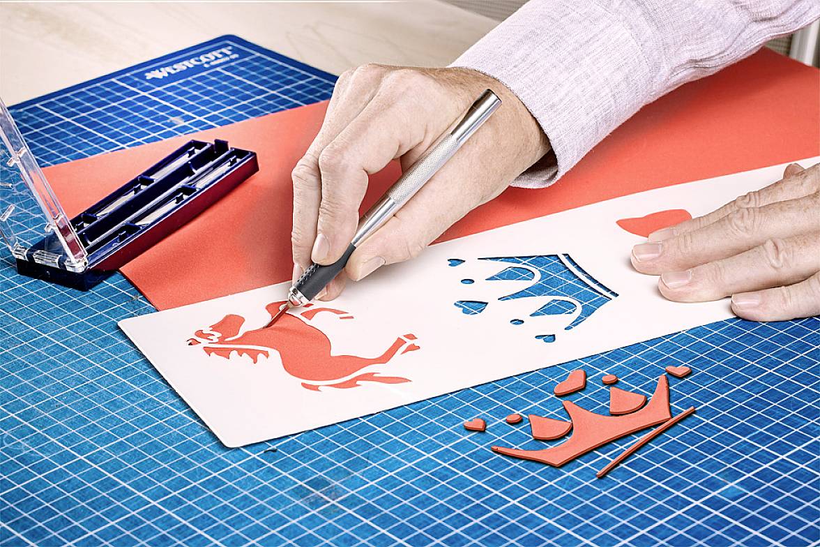 Une main découpe des formes en papier rouge, dont un cheval, une couronne et des cœurs, sur une planche à découper bleue. Des outils sont visibles.