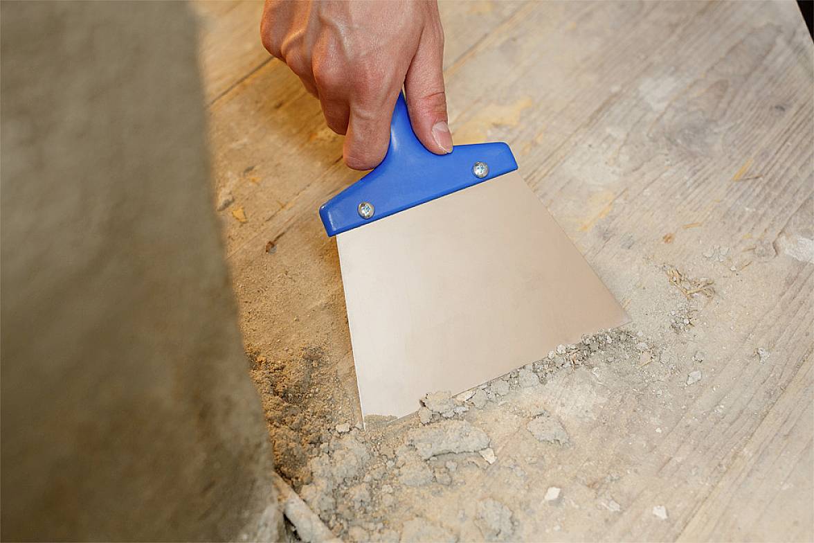 Une main tient une spatule bleue et enlève saleté et débris d'un plancher en bois.